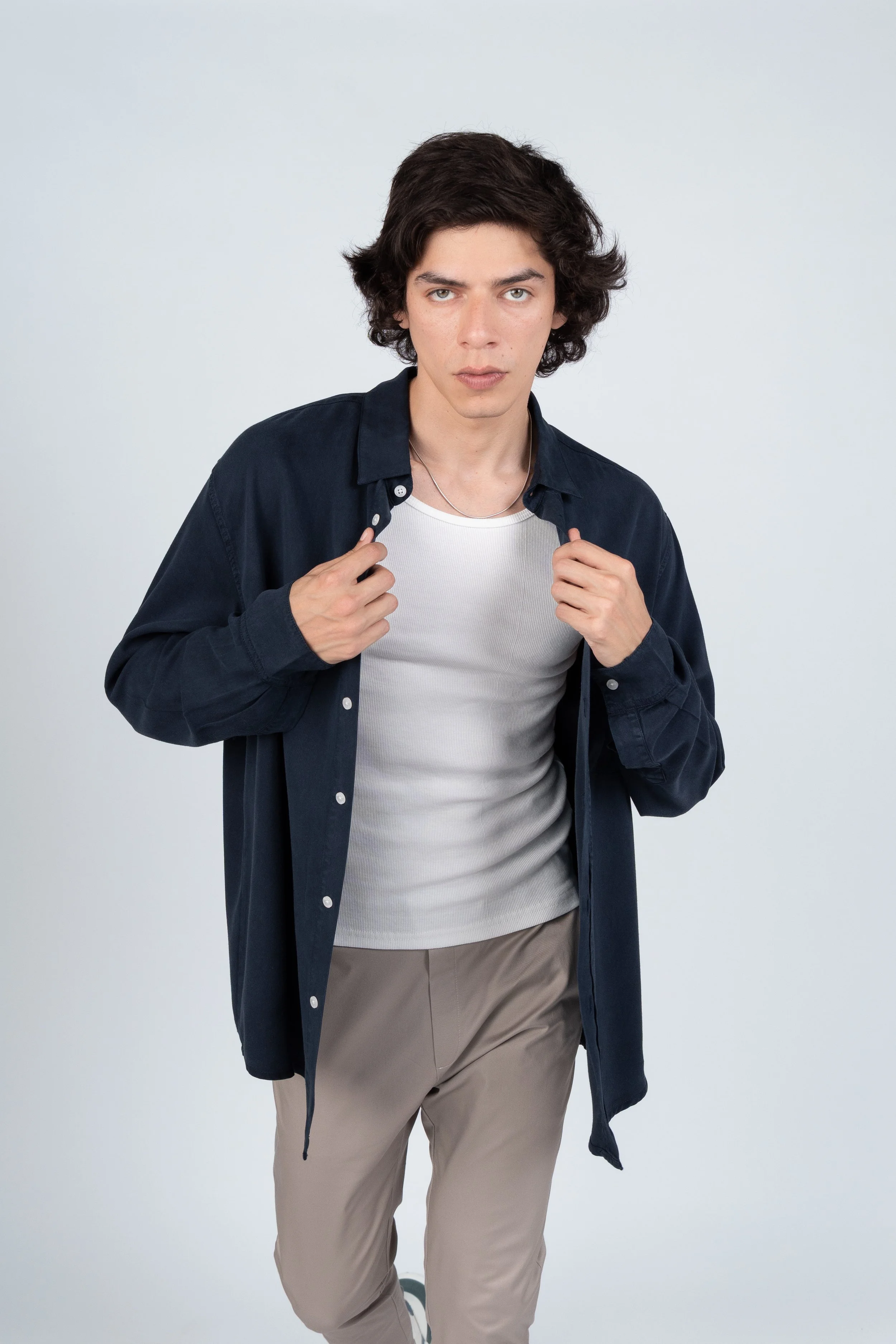 Joven con cabello rizado, camisa azul, camiseta blanca y pantalones beige, posando en un fondo gris claro.