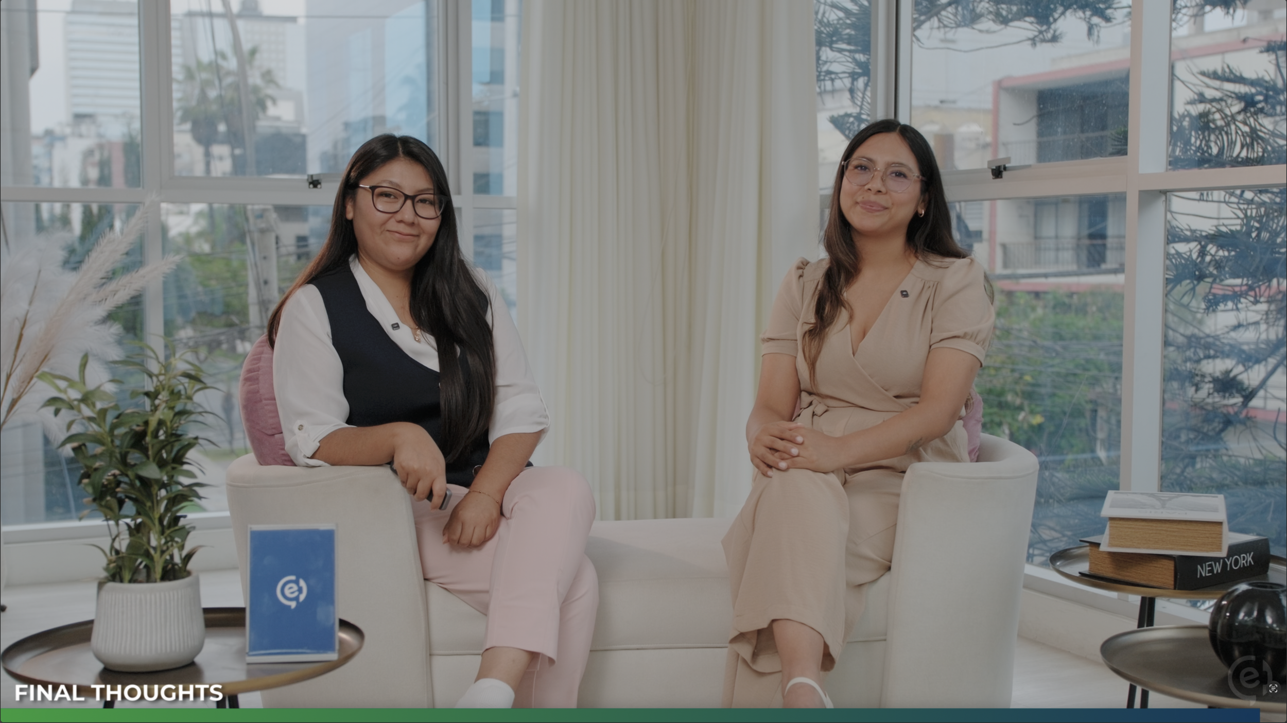 Dos mujeres sentadas en sillones blancos en una sala con ventanas grandes y vista a árboles y edificios. Una lleva gafas negras y camiseta blanca con chaqueta negra, la otra lleva gafas con marco rosado y vestido beige. Hablan hacia la cámara.