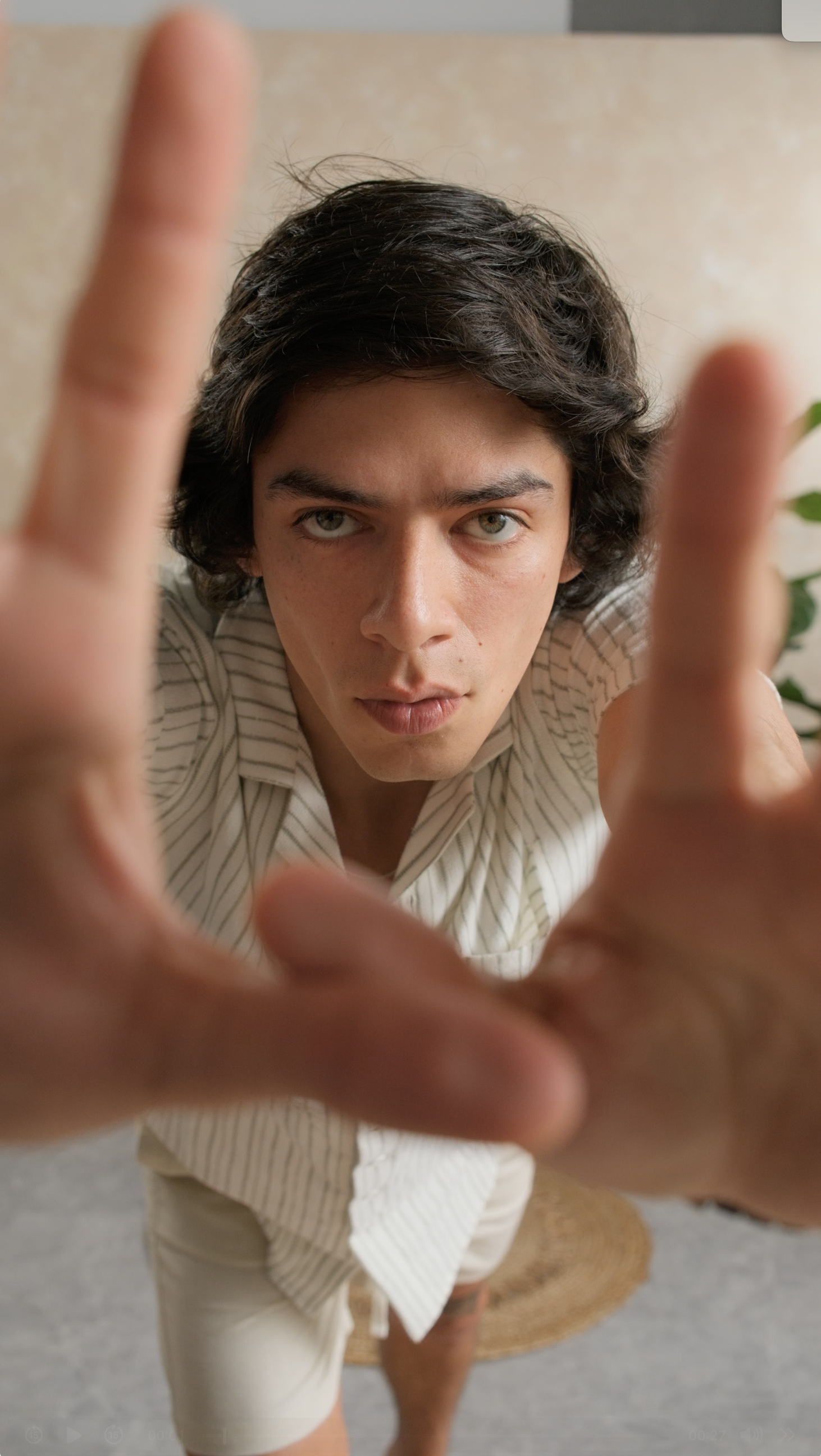 Joven con cabello oscuro y ojos claros mira hacia la cámara, con las manos cerca de la lente en primer plano, en un ambiente interior.