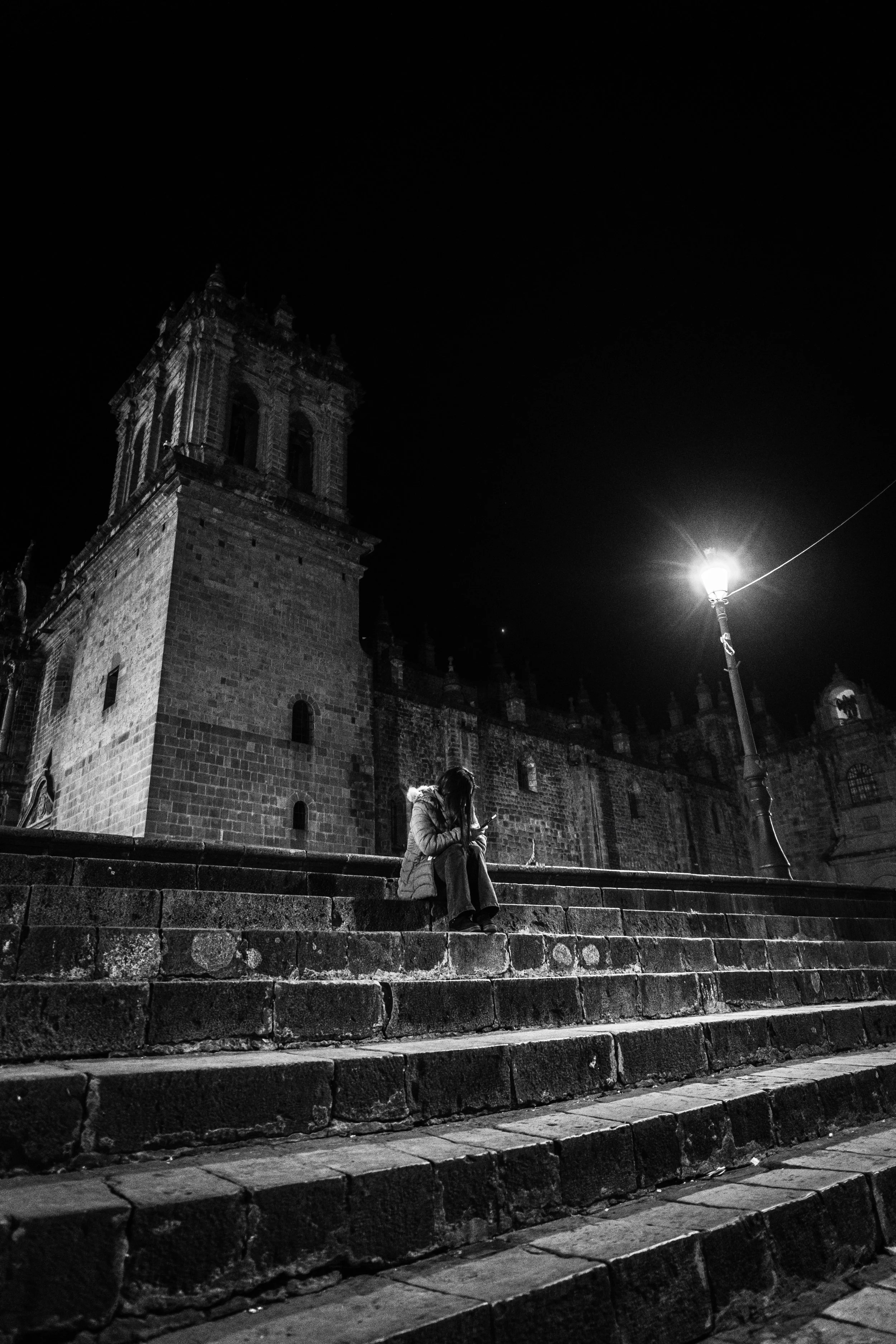 Mujer sentada en las escalinatas de una iglesia antigua en la noche, con un farol iluminando la escena y un cielo oscuro de fondo.