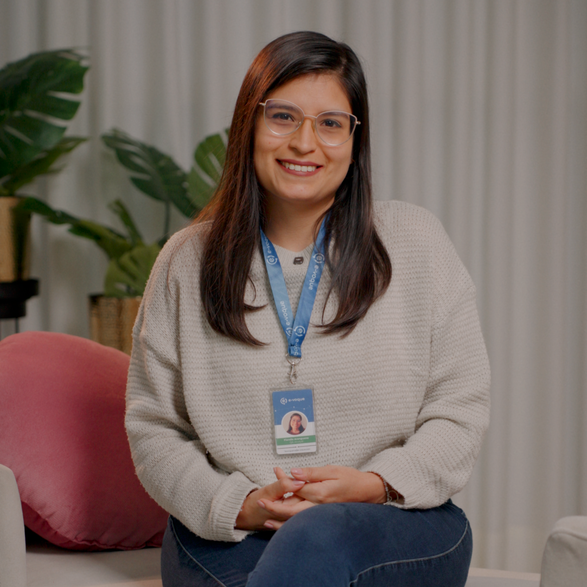 Mujer joven sonriendo, usando gafas, con una chaqueta beige y una tarjeta de identificación en el cuello, sentada en un espacio interior con cortina beige de fondo y planta verde a la izquierda.