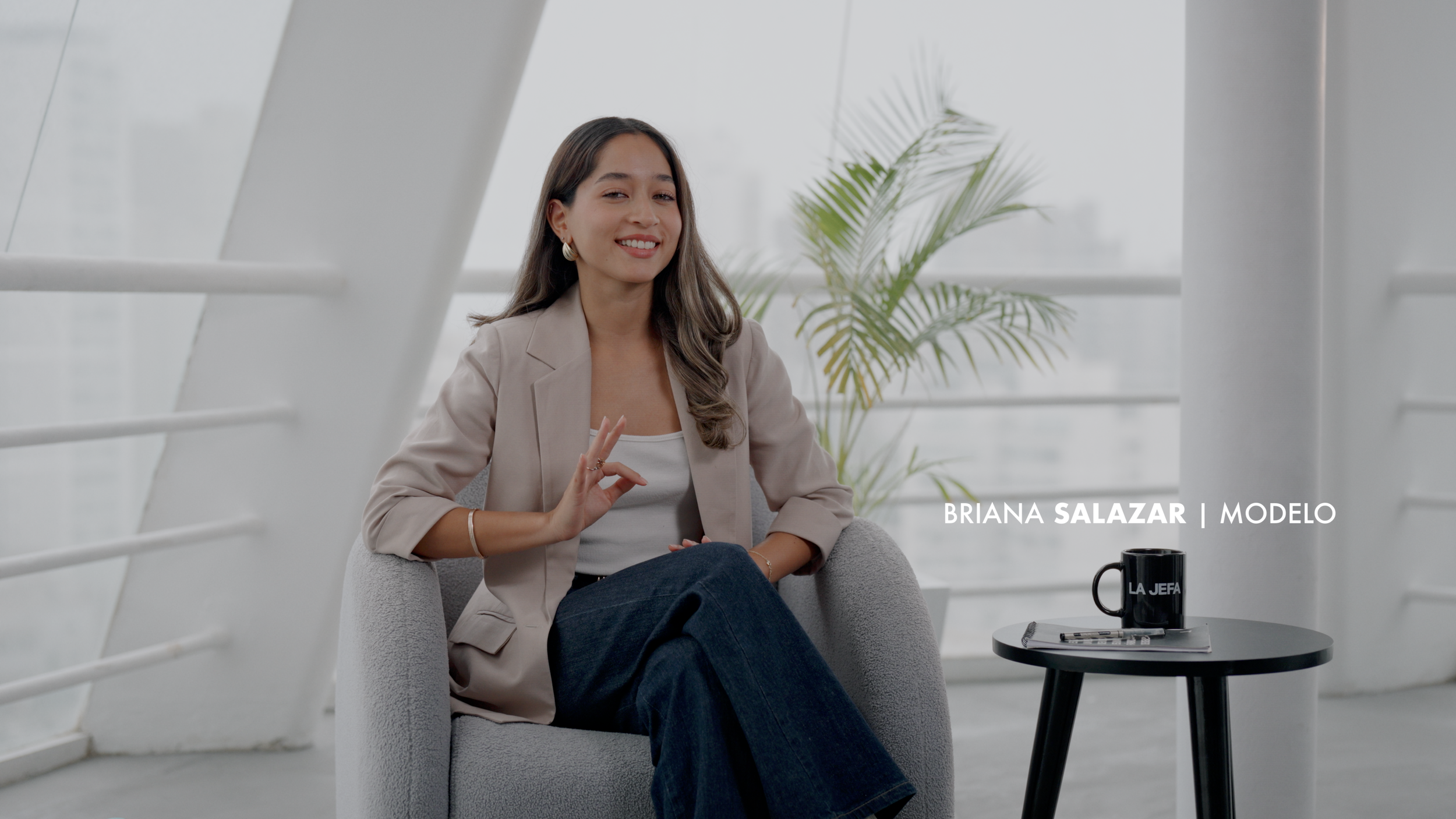 Mujer sonriendo en una entrevista, con una taza negra y algunas papeleras en una mesa, en un ambiente moderno y luminoso.