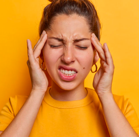 A woman with brown hair in a bun, wearing a yellow shirt, holds her head with both hands and looks uncomfortable or in pain against a yellow background, relating to tmj/tmd.