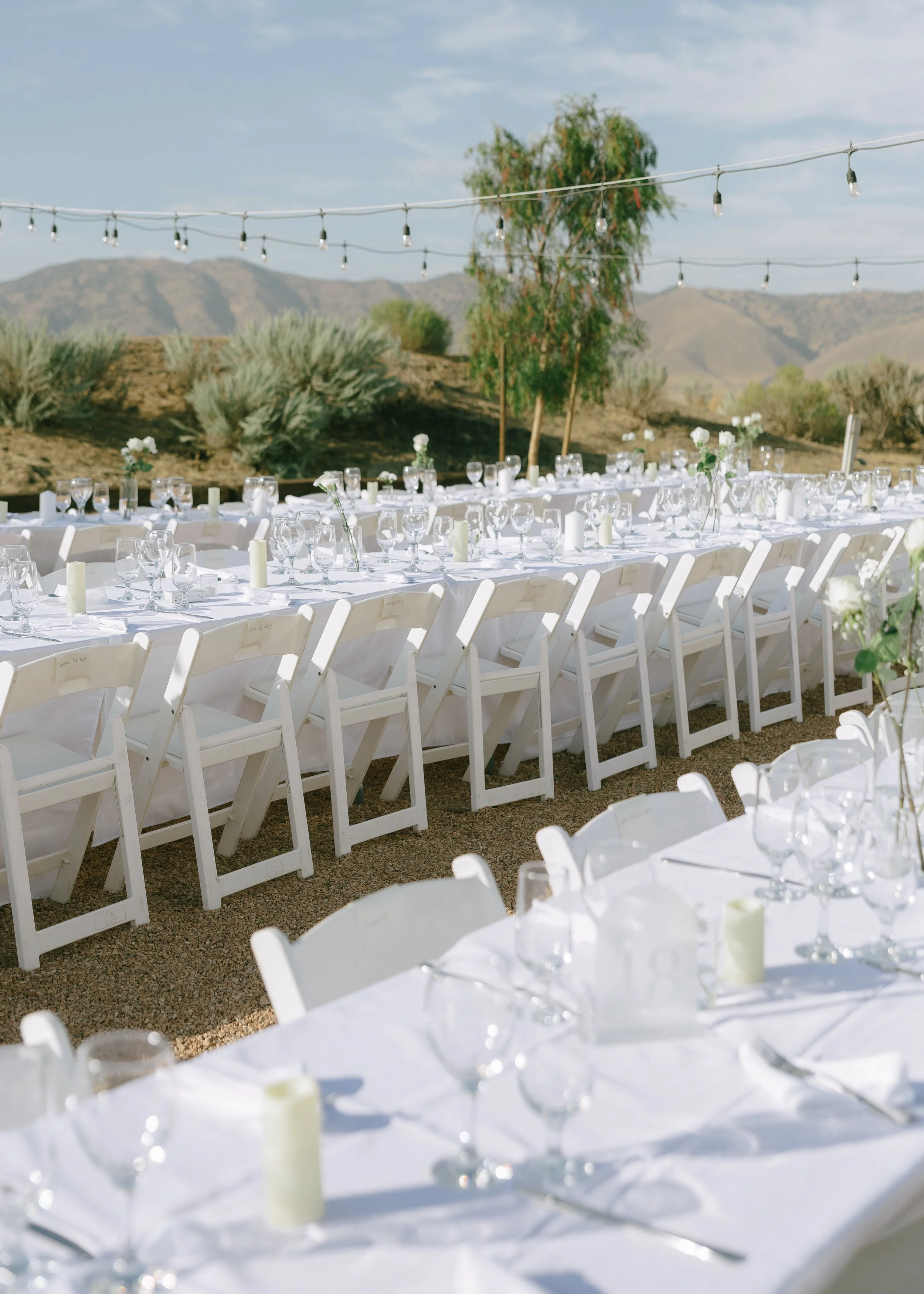 Outdoor banquet setup with white tables, chairs, glassware, and candles, against a desert landscape with mountains and string lights overhead.