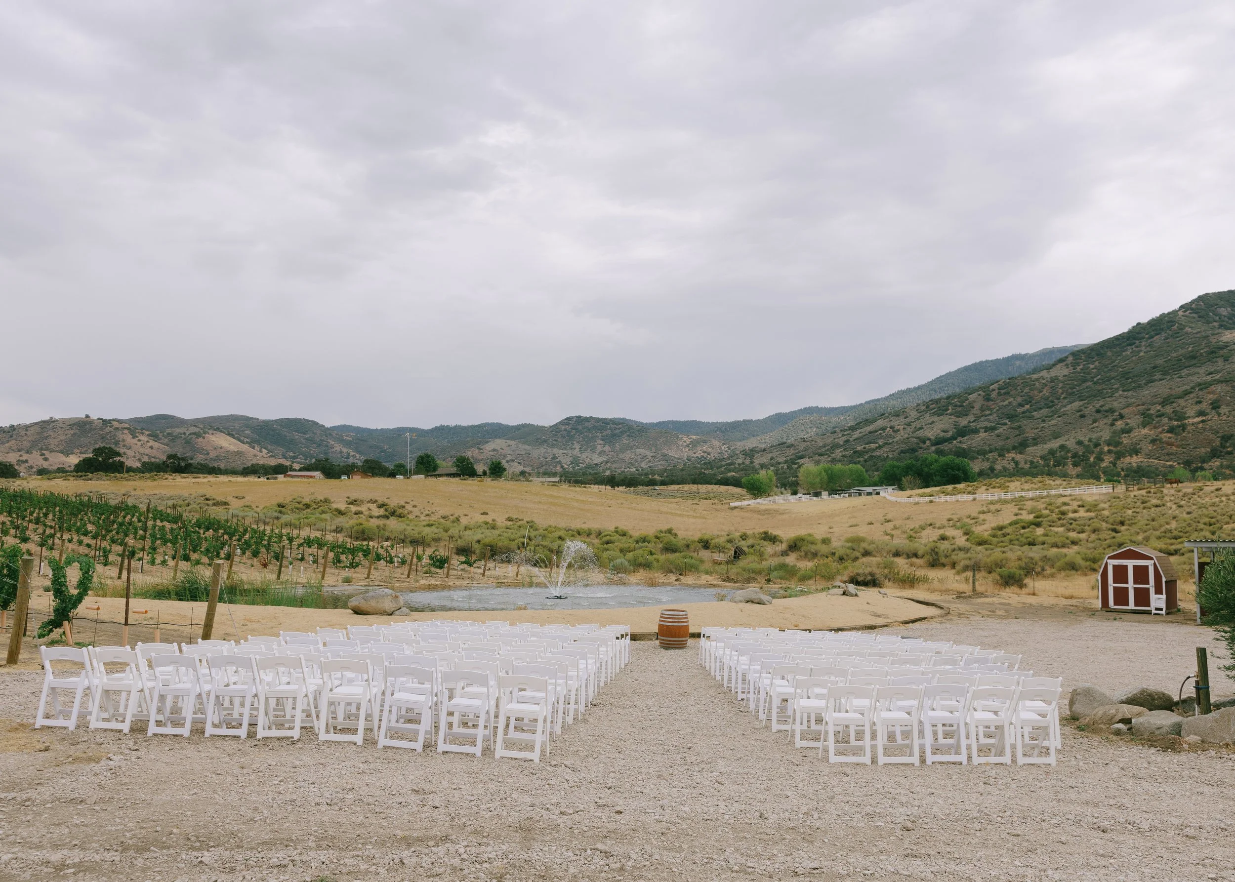Empty white chairs arranged in rows outdoors facing a small fountain, with a scenic backdrop of hills, vineyards, a pond, and a red barn under a cloudy sky.