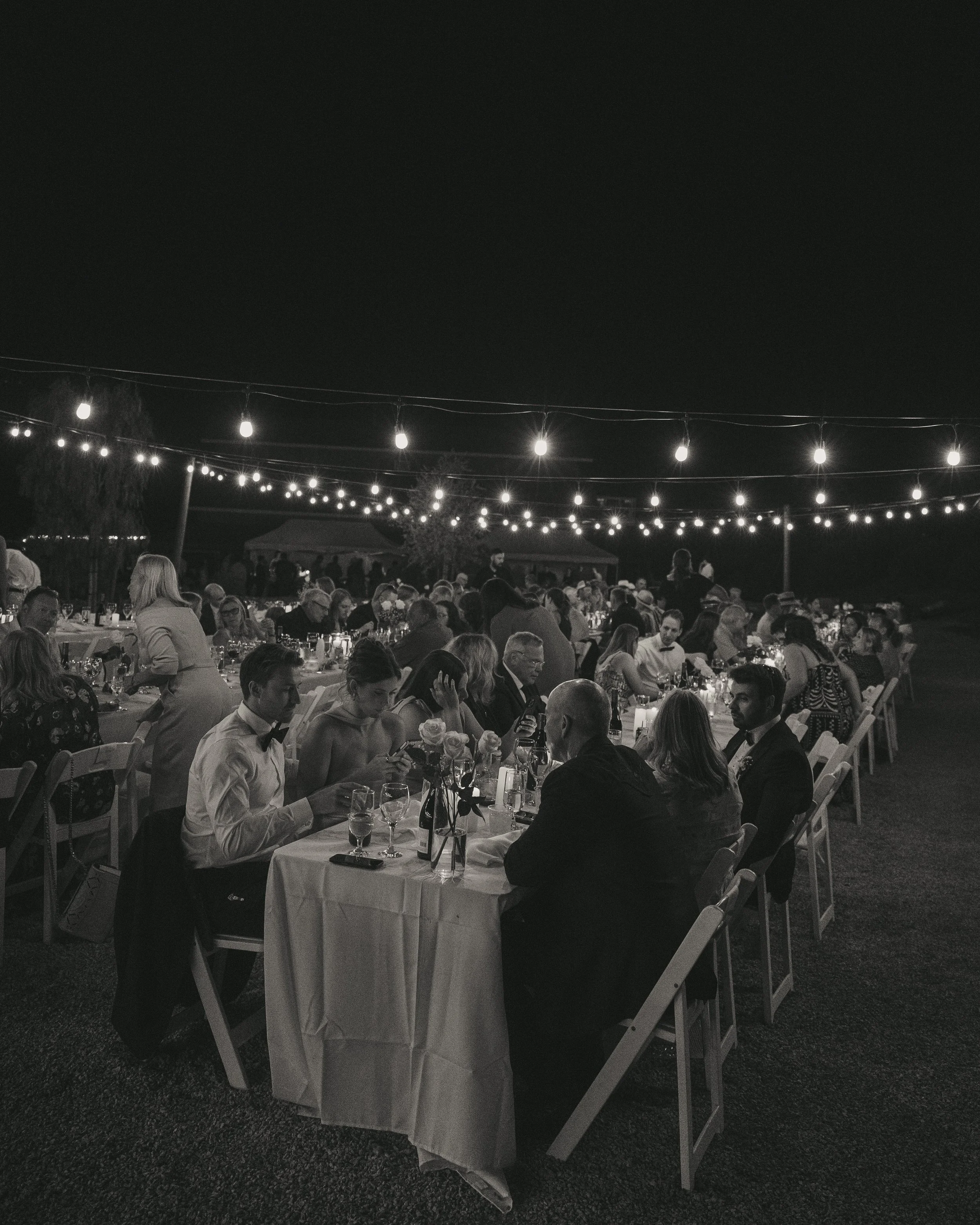 Nighttime outdoor celebration with multiple tables of formally dressed guests, string lights overhead, and a dark sky.