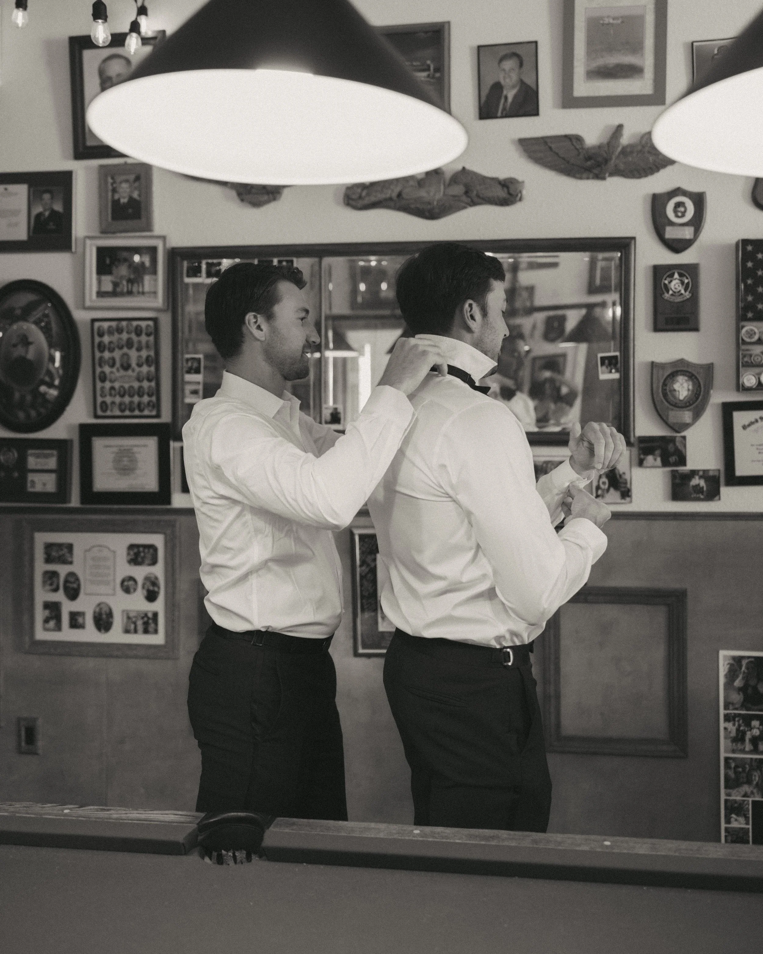 Two men in white shirts and black pants are in a room, preparing for an event. One is helping the other adjust his bow tie. The background features framed photos and plaques on the wall.
