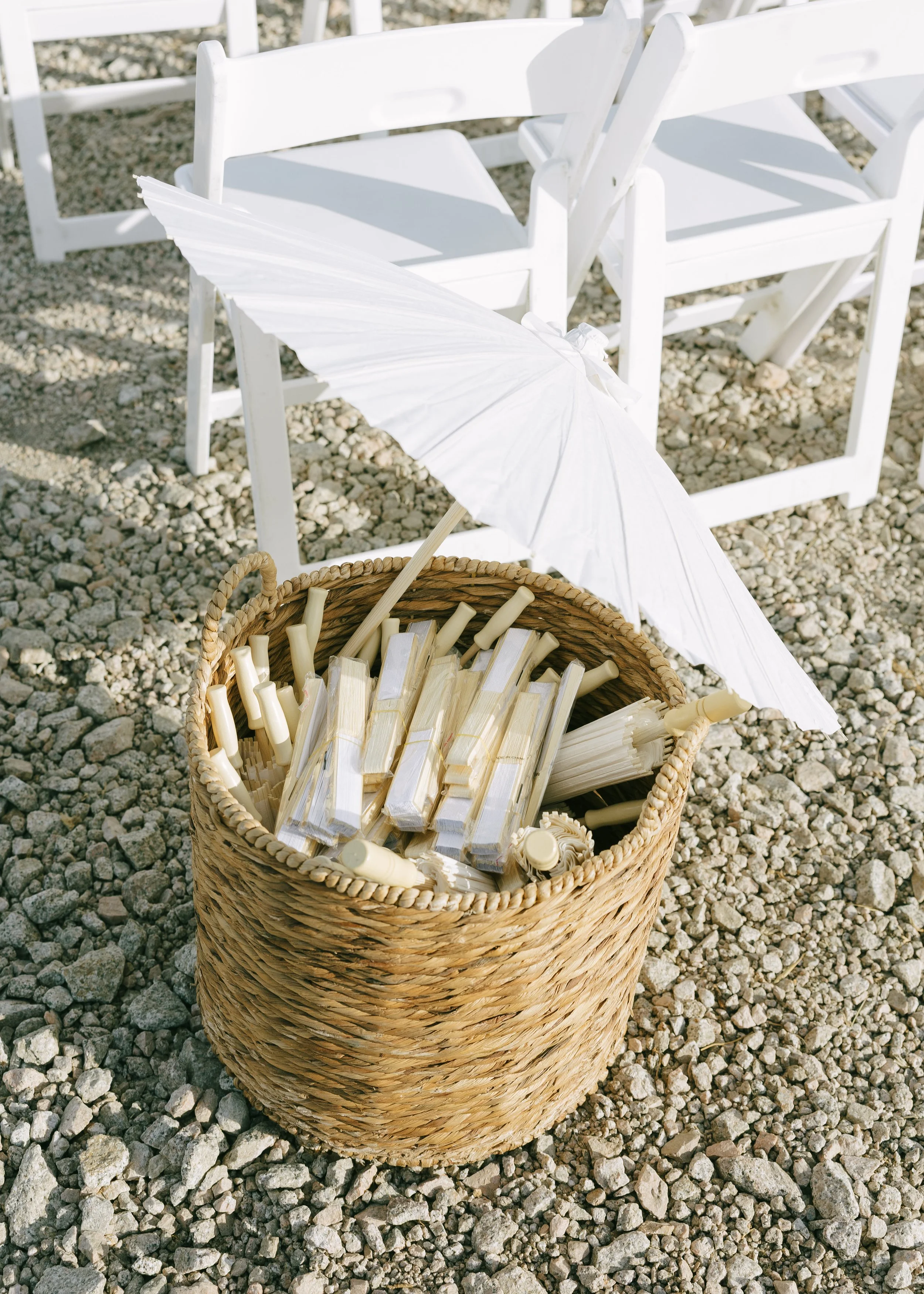 A wicker basket filled with white paper umbrellas and cocktail stirrers, with several white folding chairs on gravel ground in the background, suggesting an outdoor event setup.