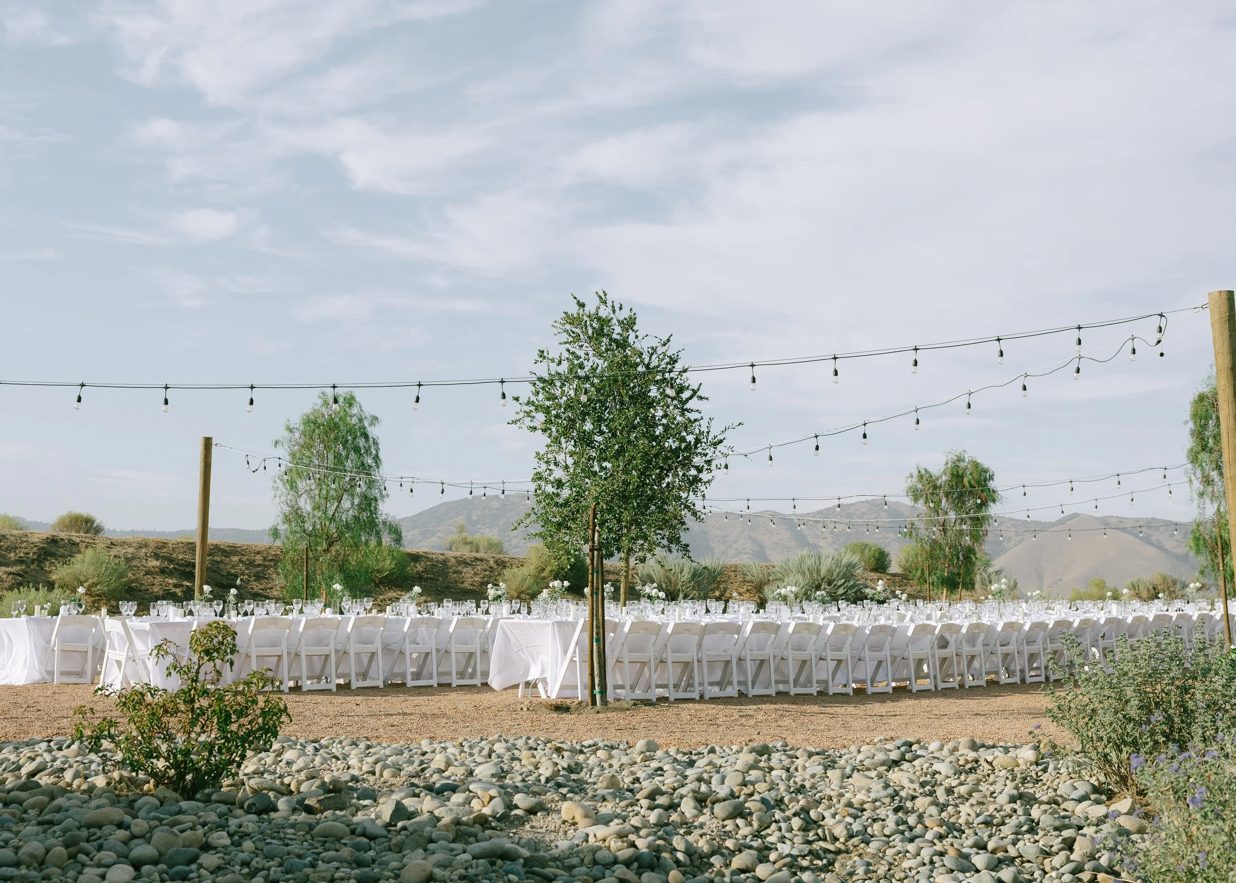 Outdoor wedding reception area with long white-draped tables, white chairs, string lights overhead, and a scenic desert landscape with mountains in the background.