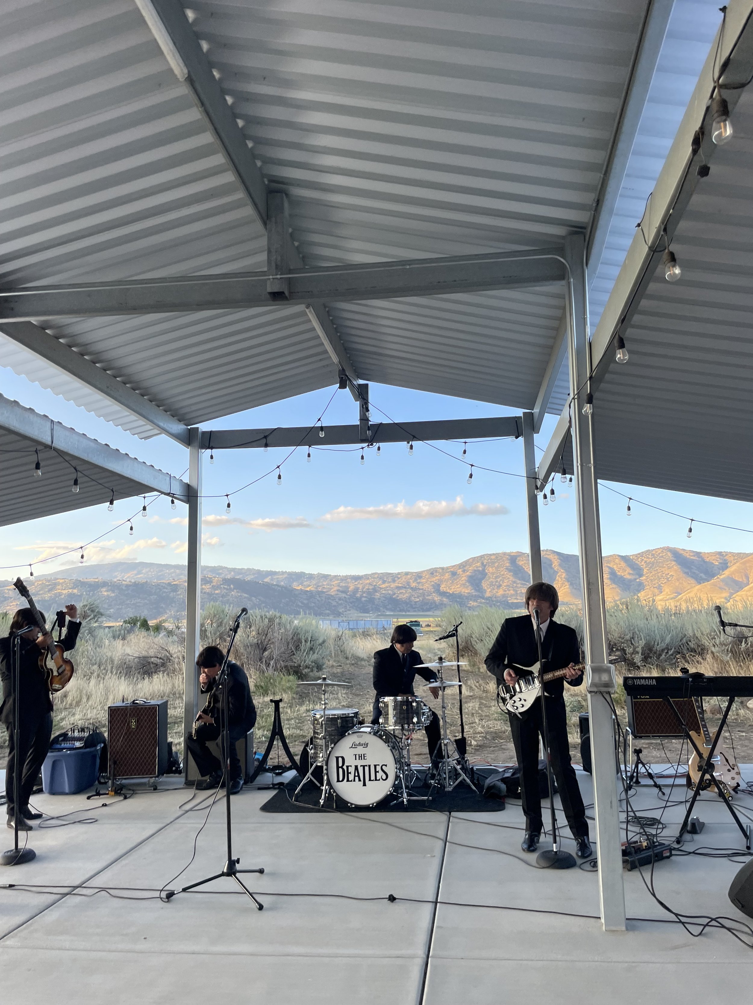 A band dressed as The Beatles performs on an outdoor stage with a mountainous landscape in the background.