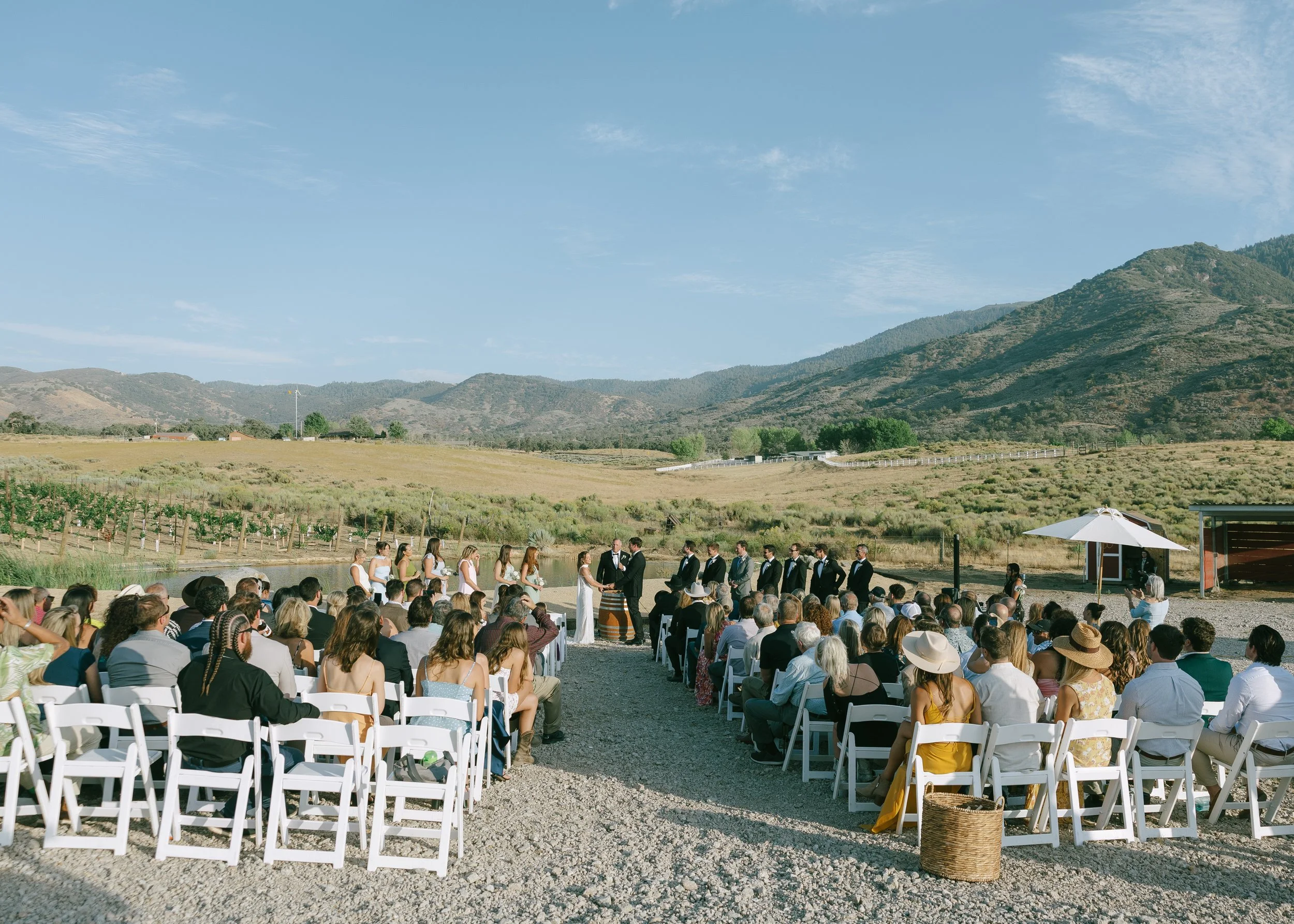 Outdoor wedding ceremony with guests seated on white chairs, marrying couple exchanging vows in front of witnesses, set against open countryside with rolling hills and mountains under a blue sky.