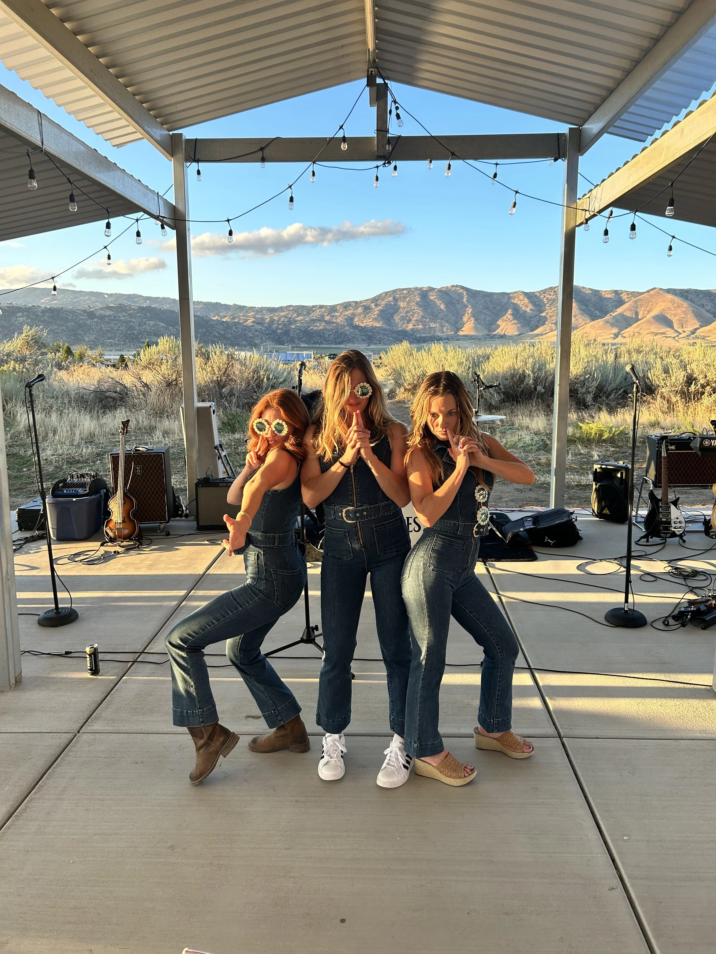 Three women in denim outfits dancing on an outdoor stage with musical instruments and mountains in the background.