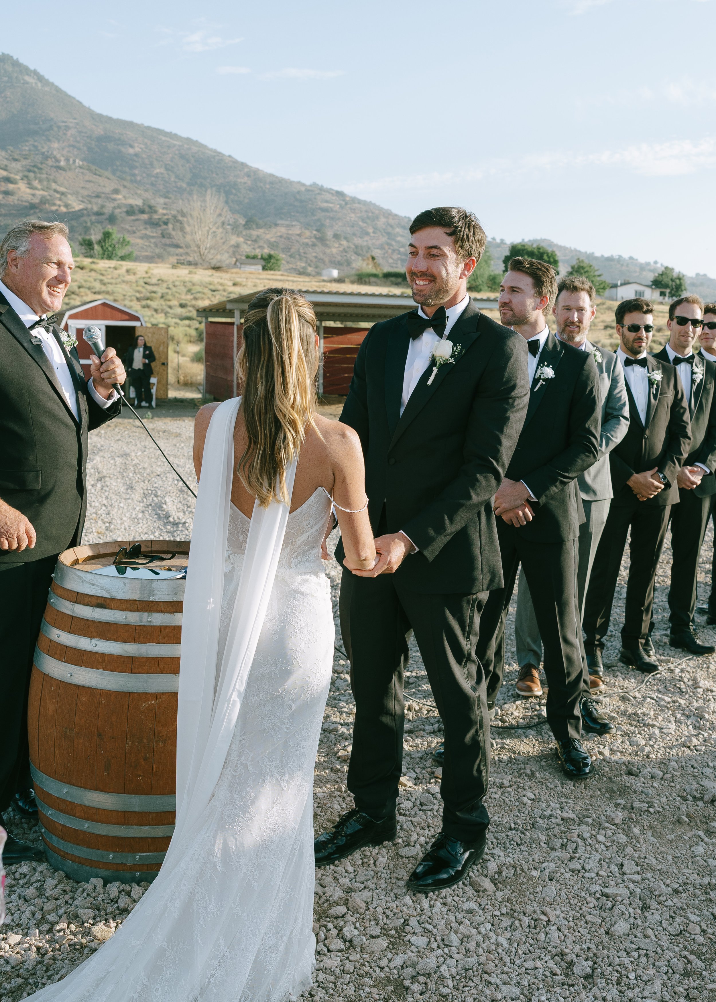 Wedding ceremony outdoors with bride and groom holding hands, officiant speaking, bridesmaids and groomsmen in the background, mountainous landscape.