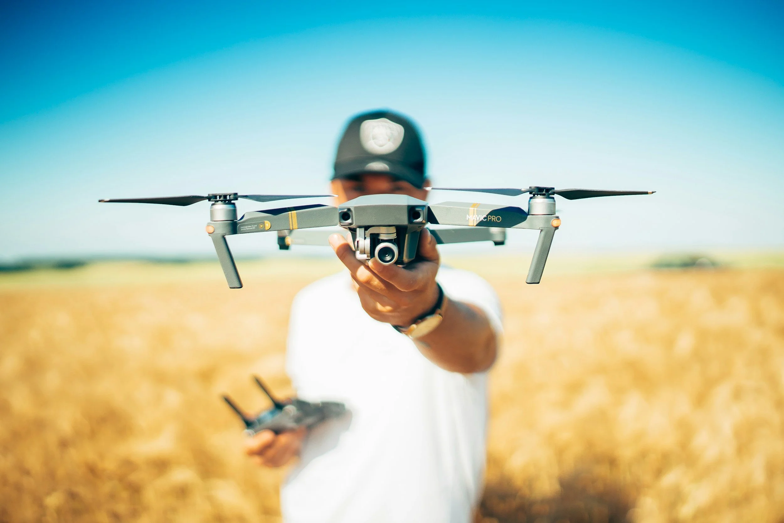 A person wearing a baseball cap flying a gray drone in a field under a clear blue sky. The person is holding a remote control in their left hand and the drone in their right hand, aiming it toward the camera.