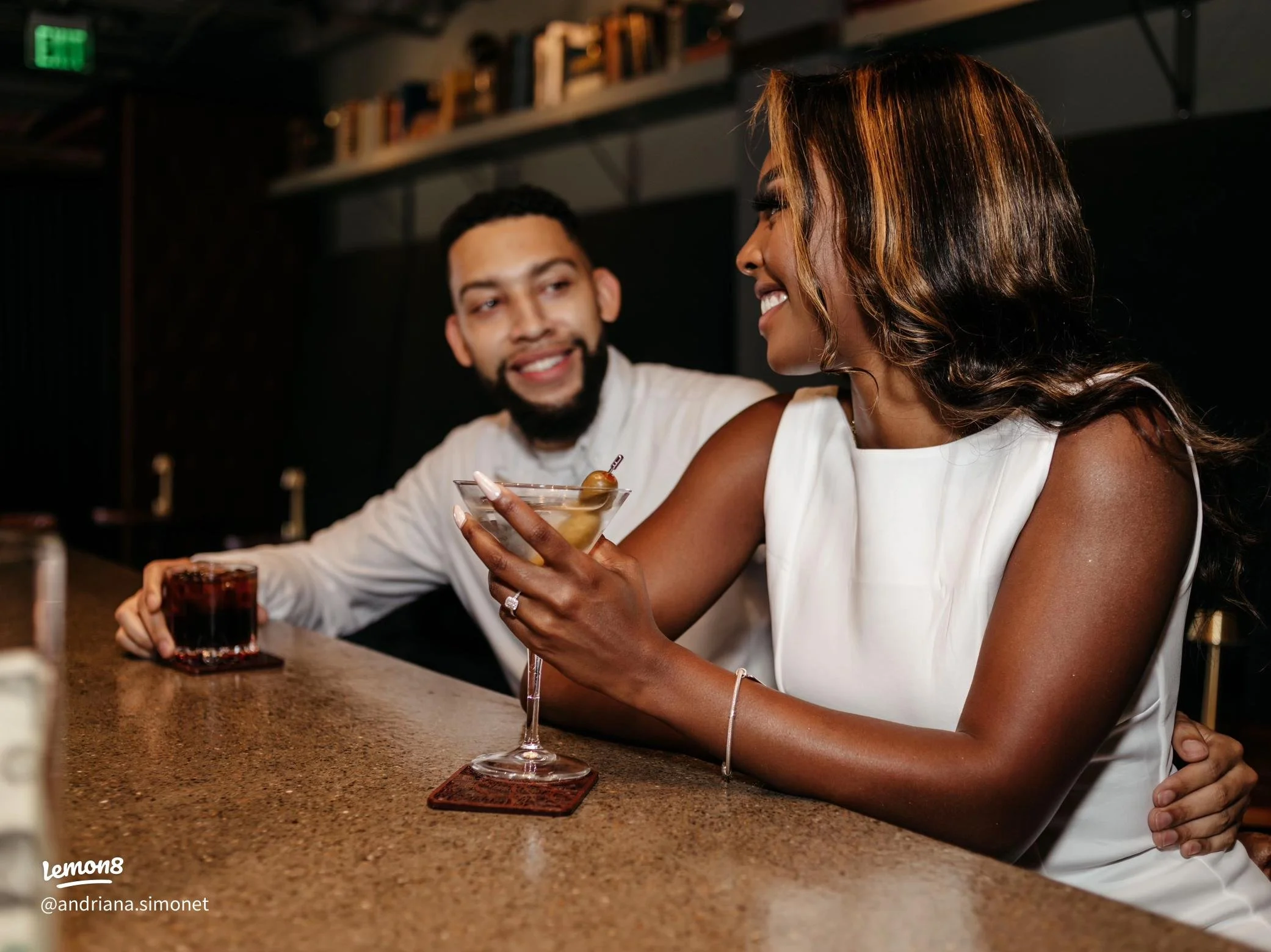 A man and woman sitting at a bar, smiling and enjoying drinks. The woman is holding a cocktail glass with an olive garnish, and the man has a glass of dark beverage. They are engaging in conversation.