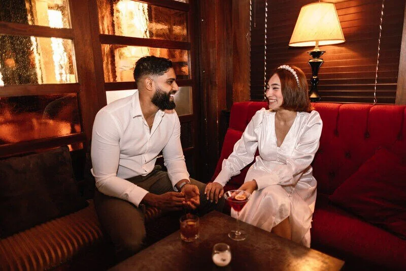 A man and woman sitting on a red sofa in a cozy dimly lit room, holding hands and smiling at each other, with drinks on the table in front of them.