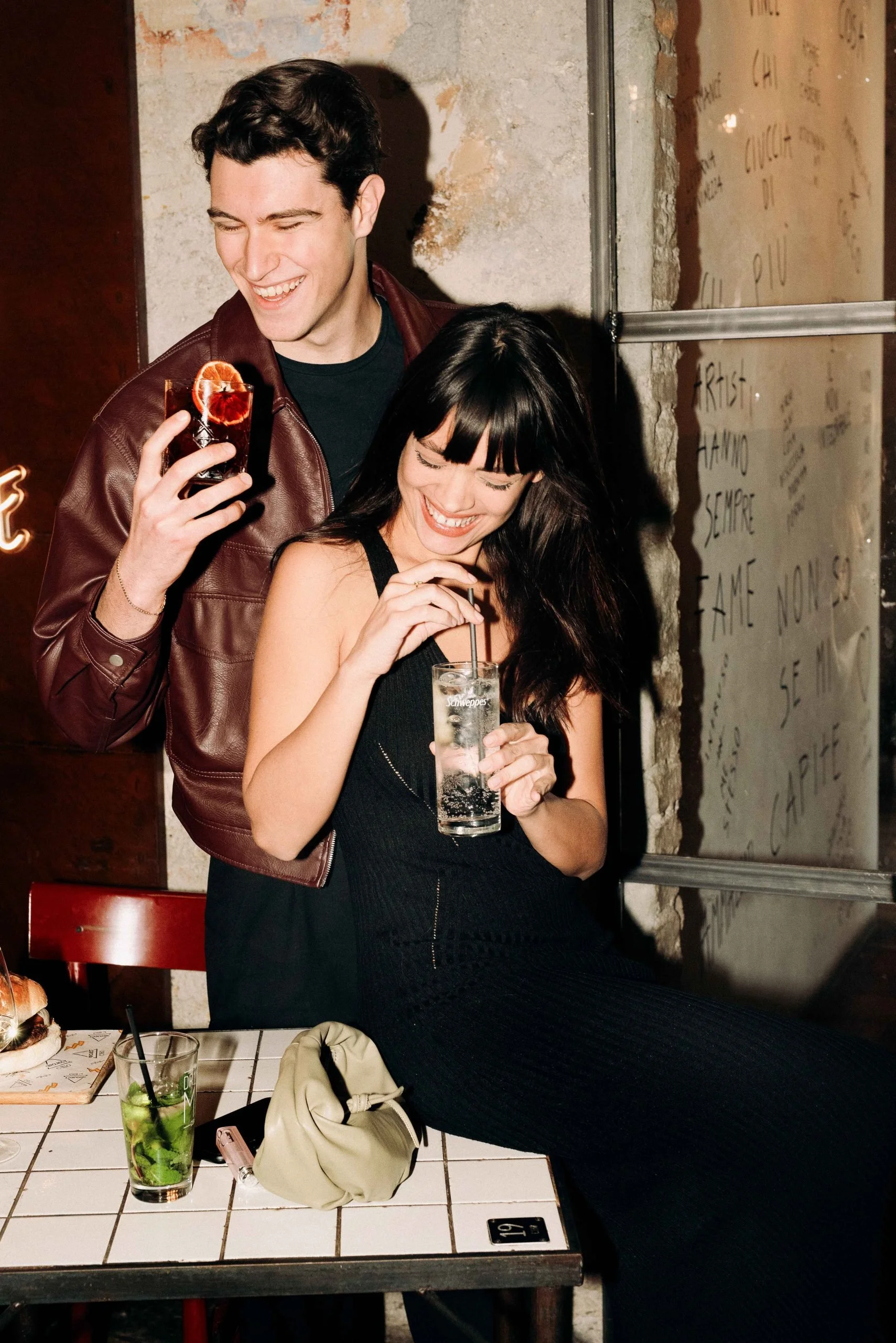 A man and woman at a bar or restaurant laughing and holding drinks. The man has dark hair and is wearing a red leather jacket, while the woman has long dark hair and is wearing a black dress. There are two drinks on the table: a mojito and a clear soda. The setting has a rustic wall with visible bricks and writing.