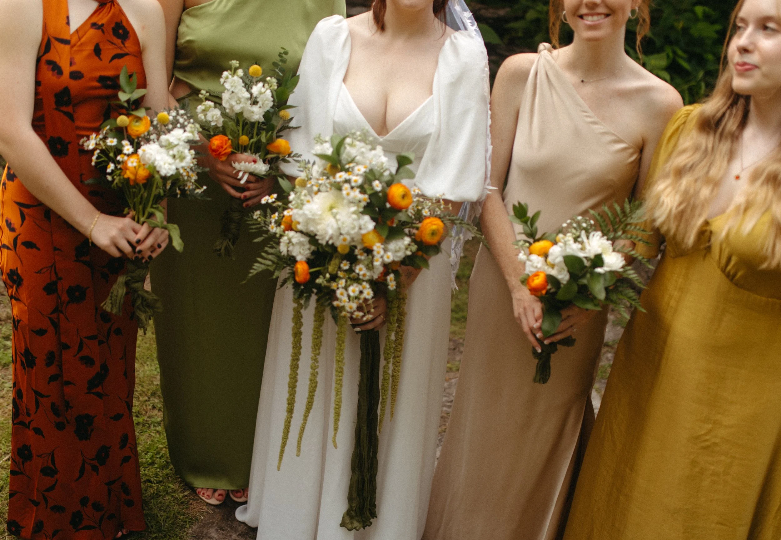 Bridal party holding bouquets at a wedding, including women in colorful dresses and an off-white gown, standing outdoors.