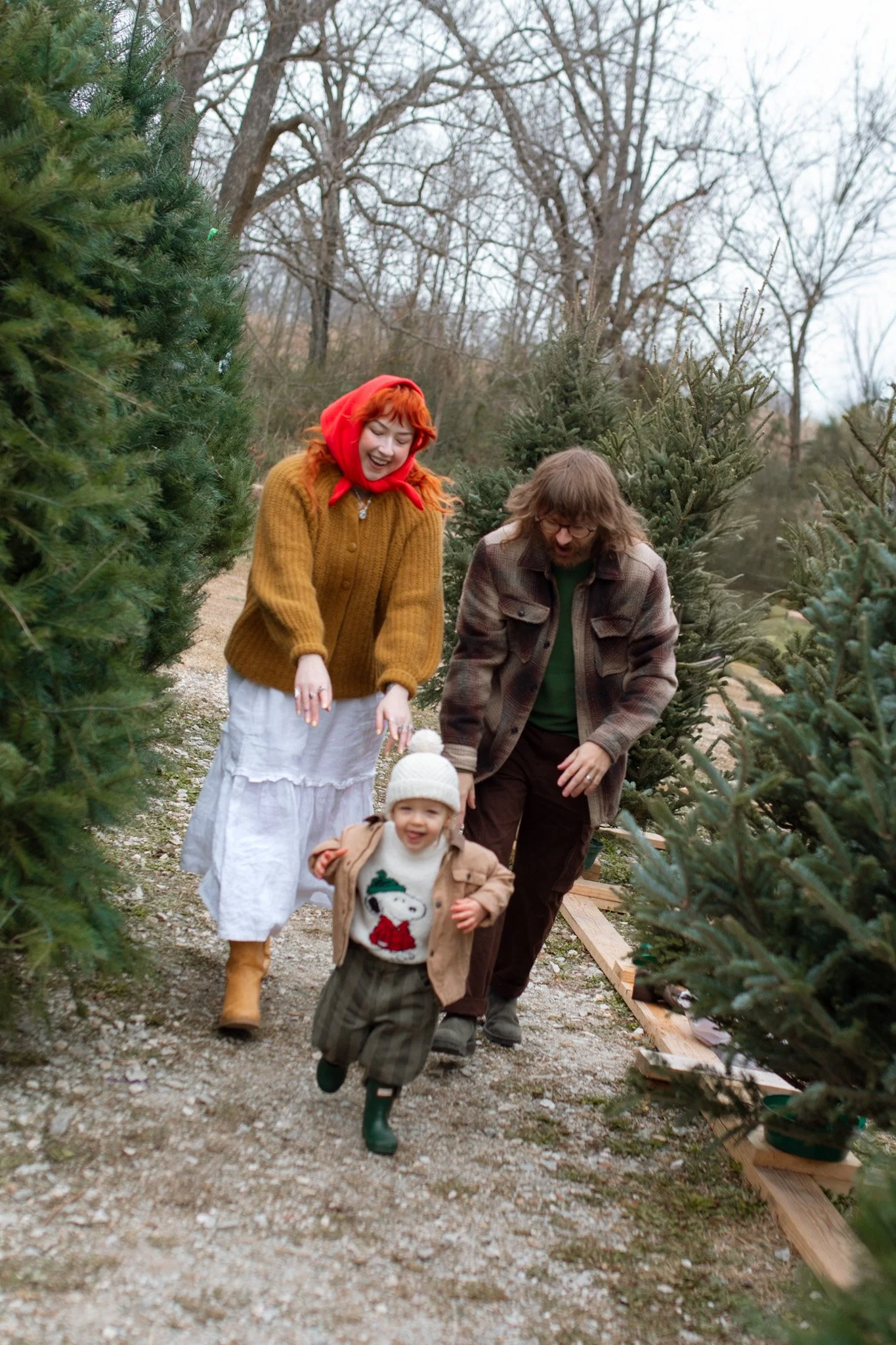 A family of three, including a toddler, joyfully walking through a Christmas tree farm with decorated evergreen trees.