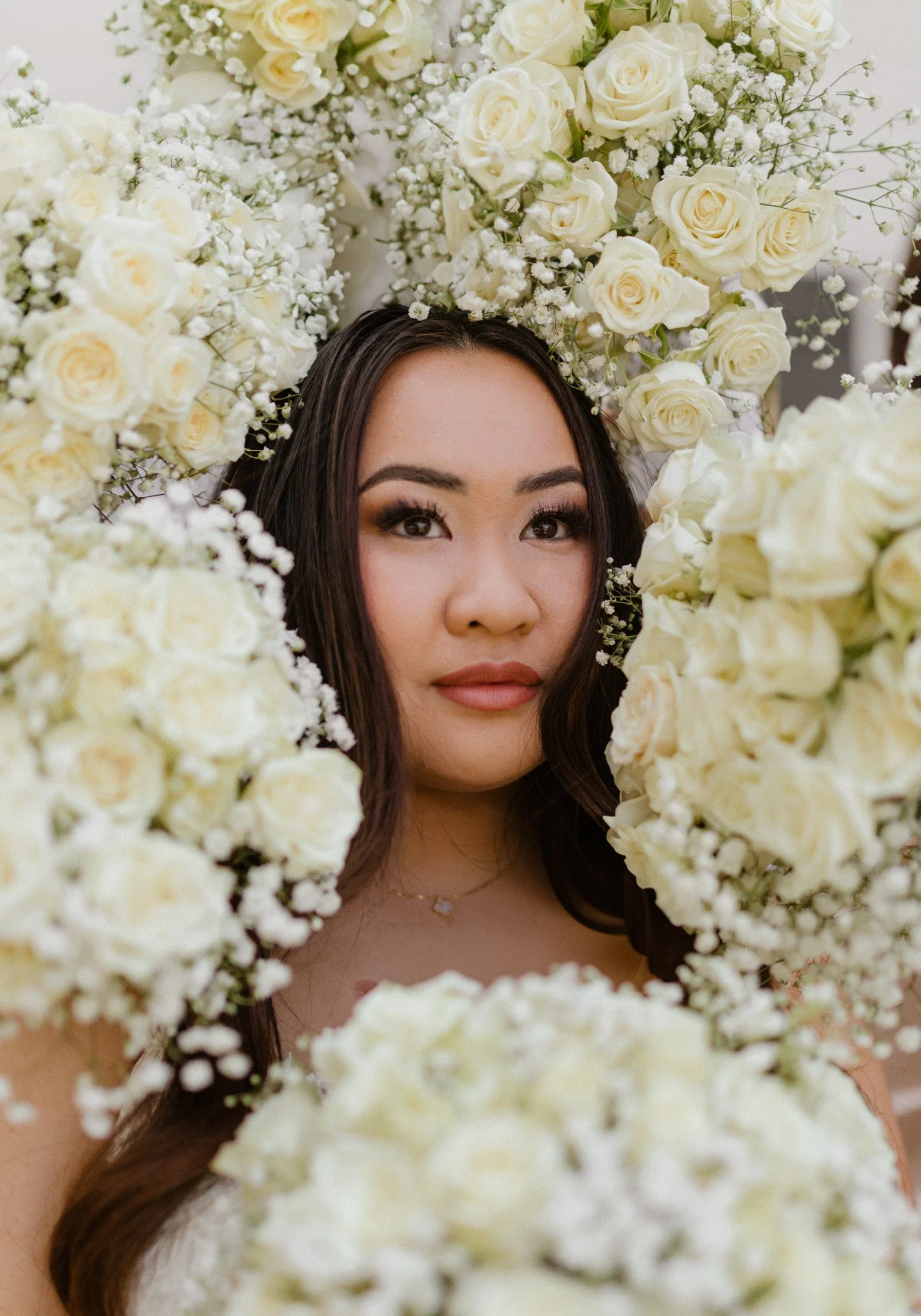 A woman with long dark hair and makeup, surrounded by a floral arrangement of white roses and baby's breath, gazing at the camera.