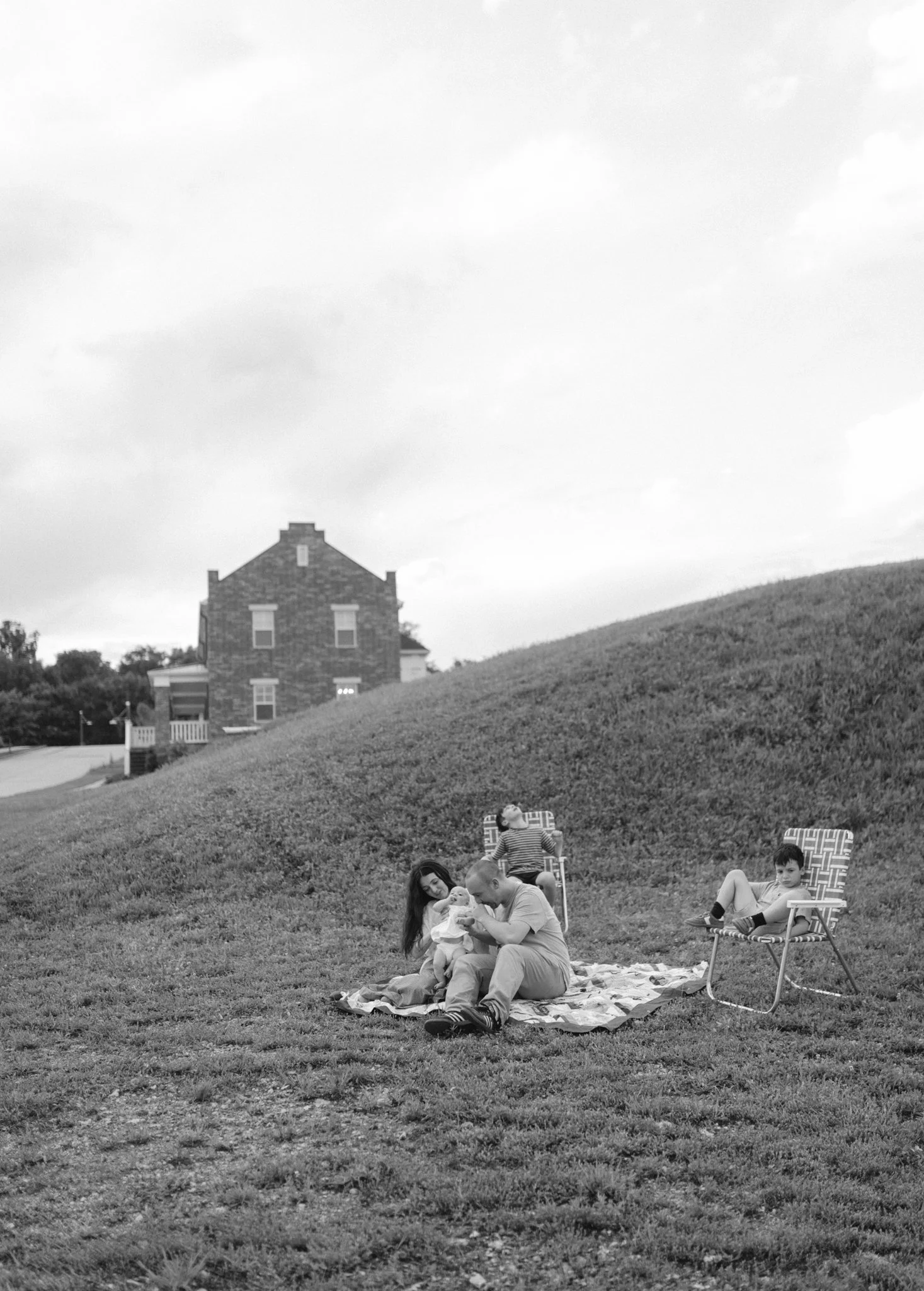 A family sitting on a blanket on the grass in a park, with two children in chairs nearby and a house in the background.