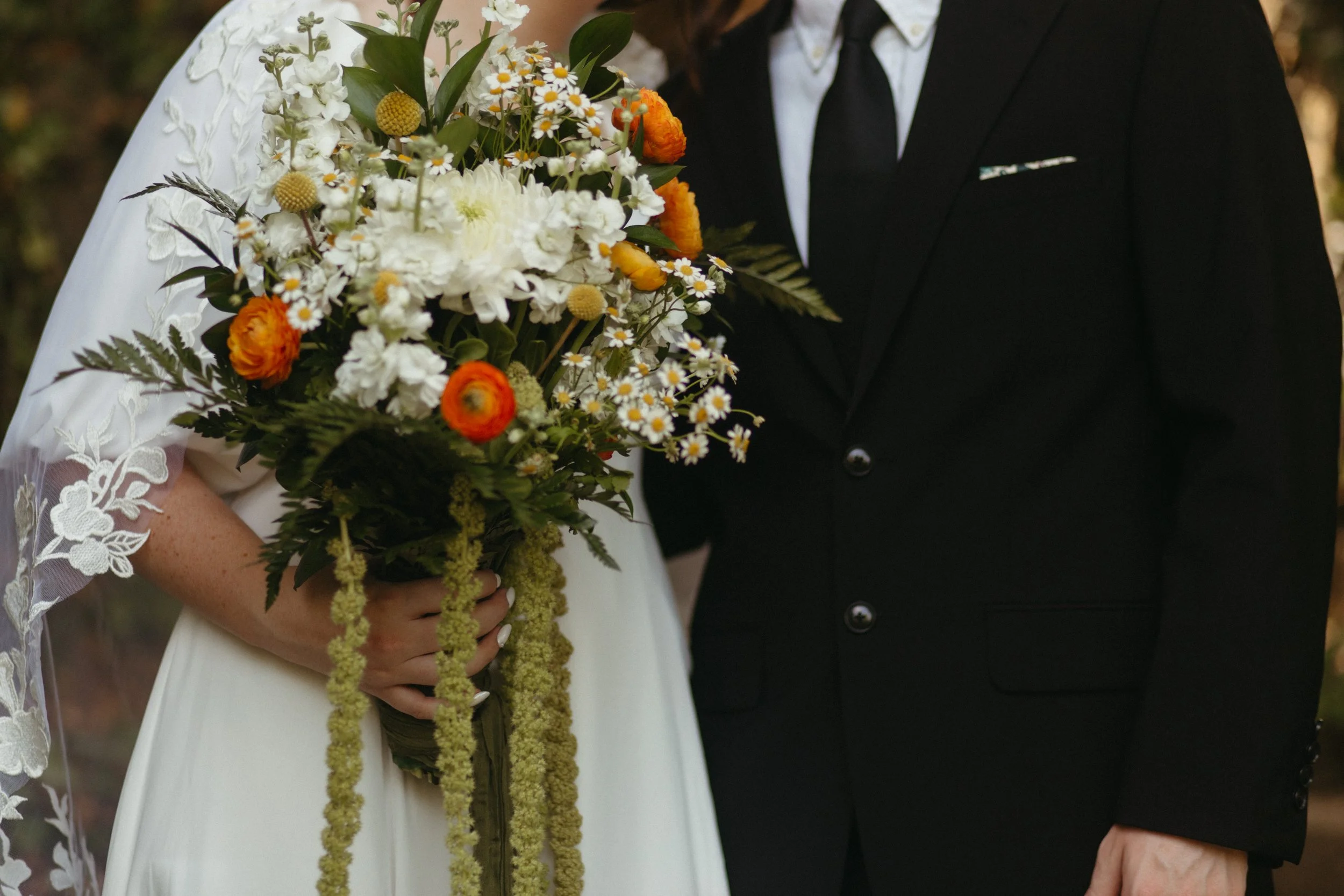 A bride holding a bouquet of white, orange, and yellow flowers, with a groom wearing a black suit standing beside her.