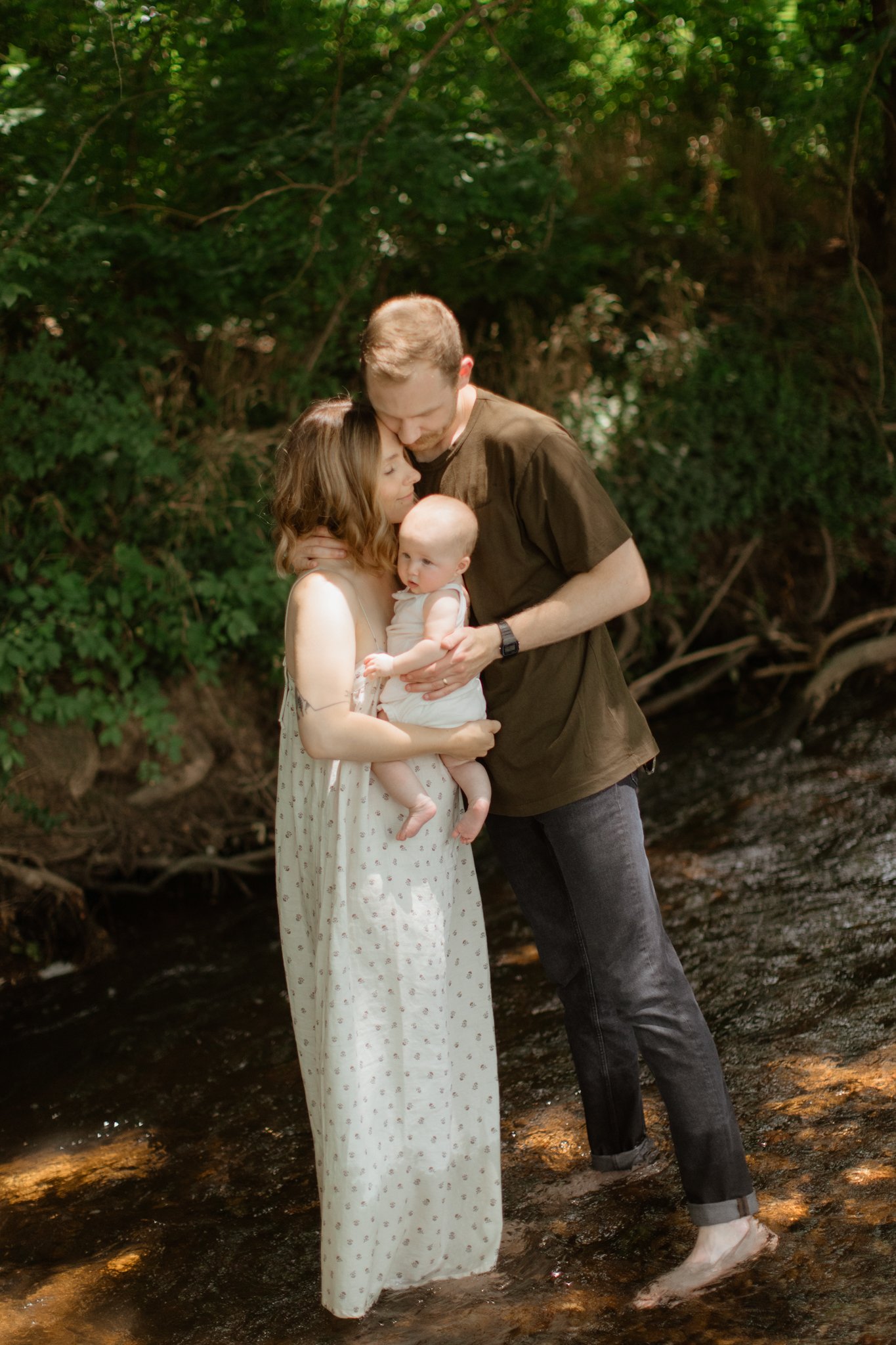 A family of three standing in a creek surrounded by green trees, with the mother holding a small baby and the father leaning in close, all barefoot and sharing a tender moment.
