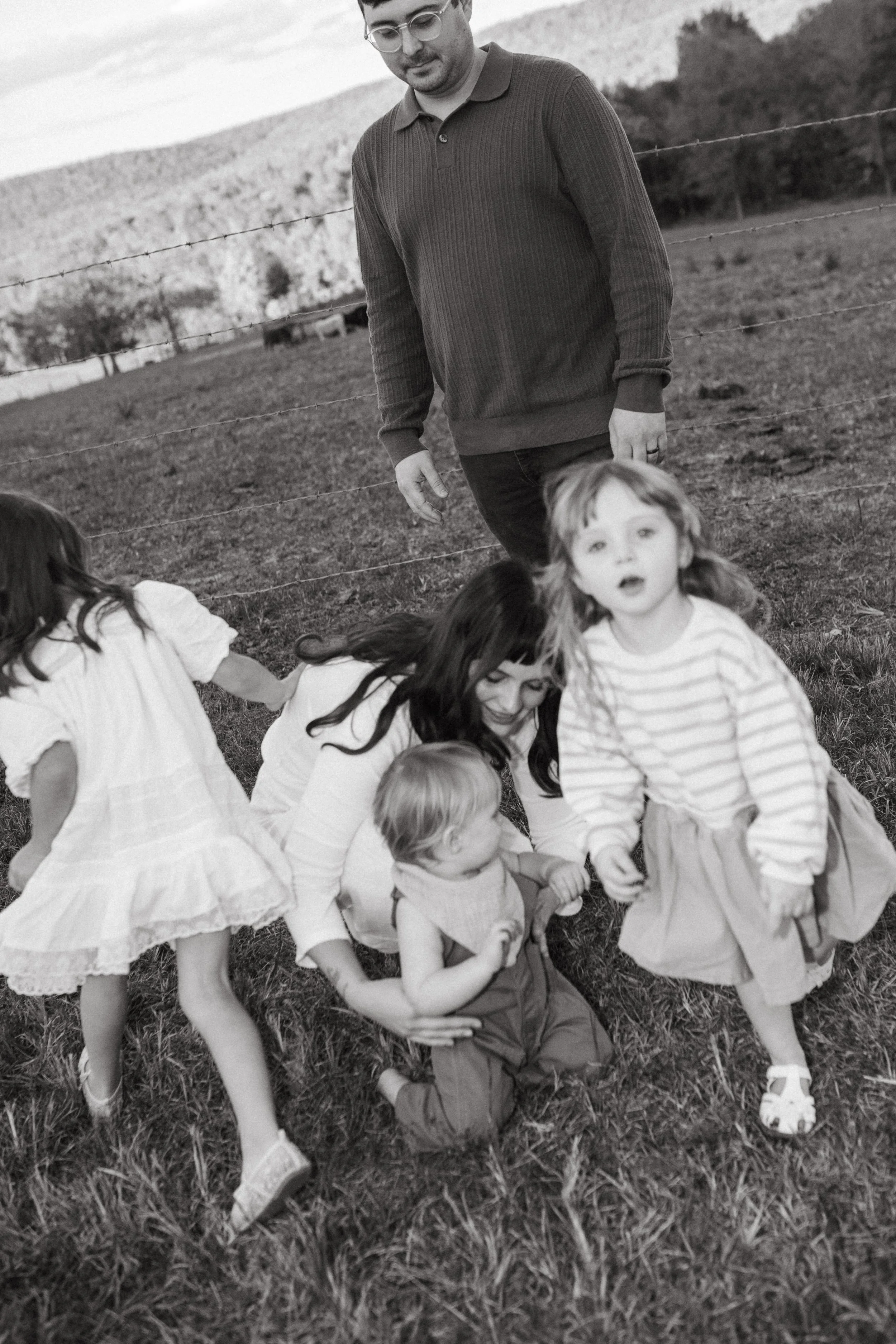 A group of children and an adult outdoors in a grassy field, with a wire fence and hills in the background.