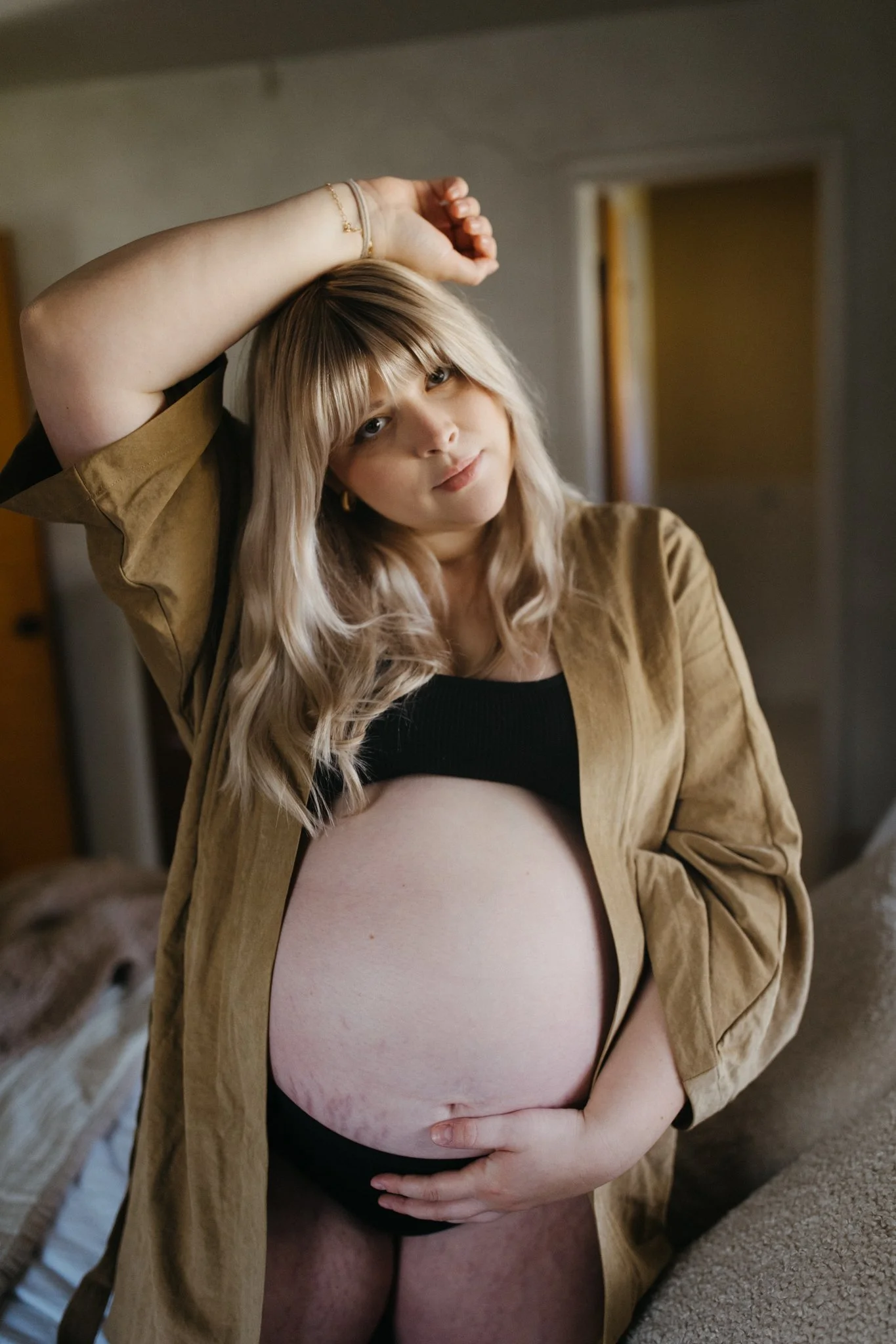 A pregnant woman with blonde hair, wearing a black crop top and a beige jacket, is standing indoors with one arm raised and the other hand on her belly, looking into the camera.