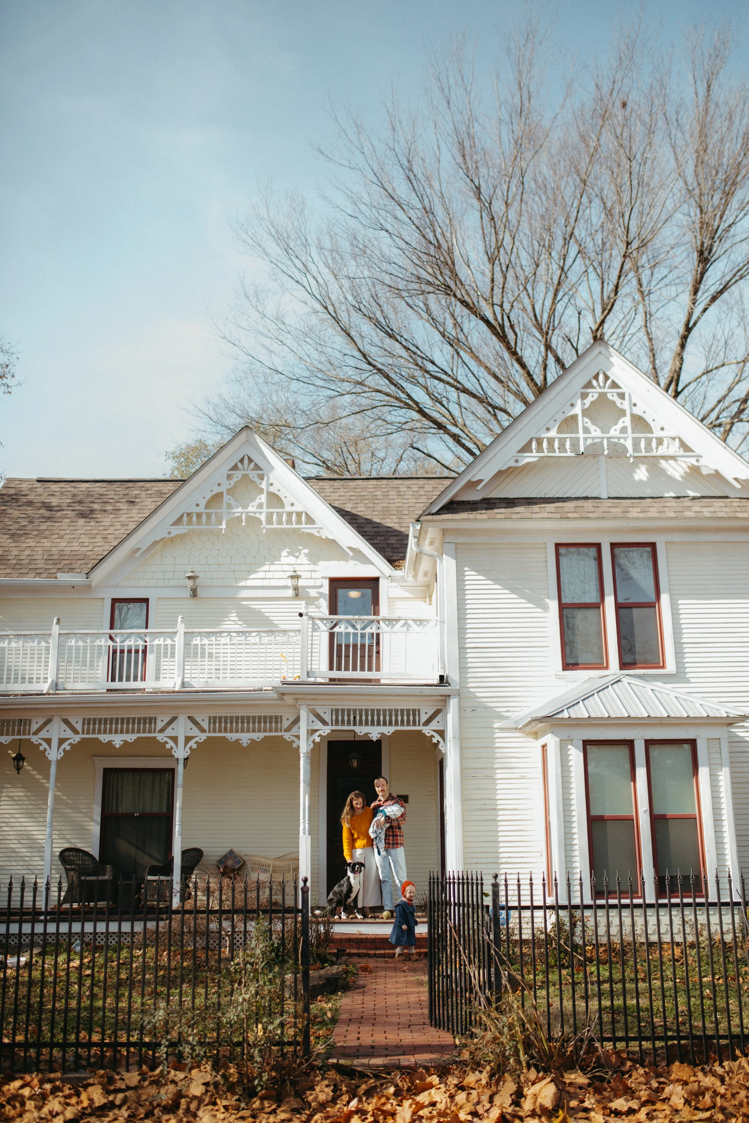 A family standing on the porch of a large white Victorian house with a small child and a dog, autumn leaves in the yard.