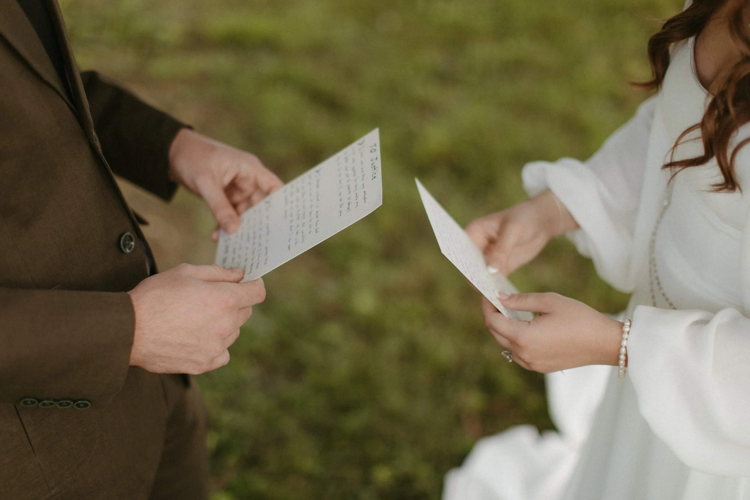 A couple exchanging vows or reading vows on their wedding day outdoors, with the focus on their hands and vows, and the background is blurred.