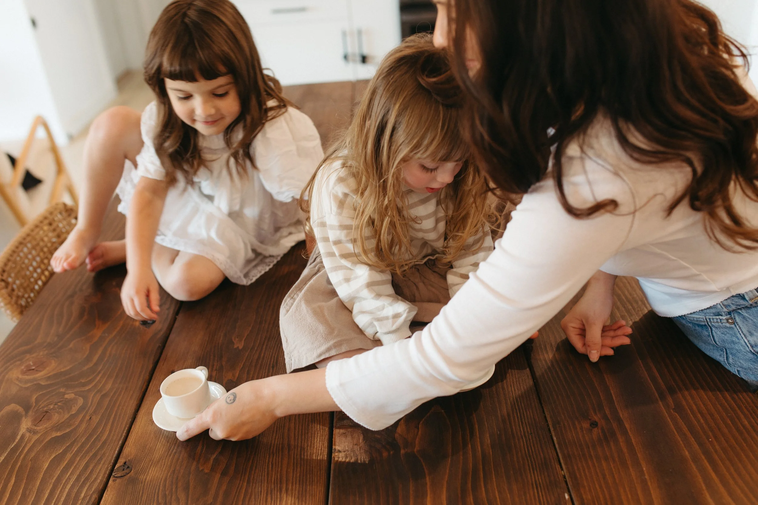 A woman and two young girls on a wooden table, with the woman helping one girl who is sitting cross-legged, while the other girl watches, in a bright room.