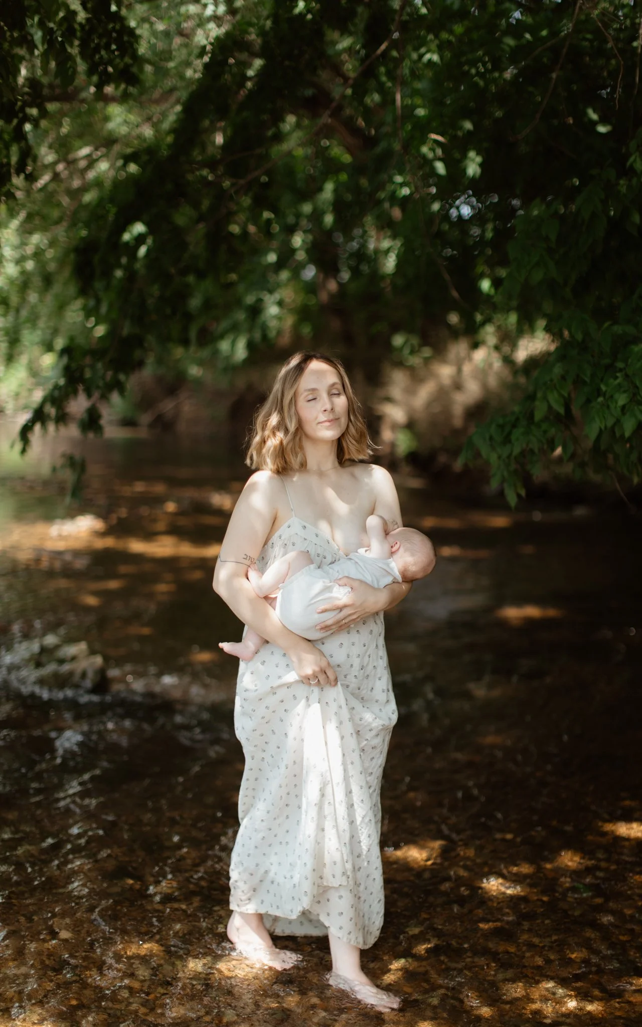 A woman stands in a shallow stream holding a baby, with her eyes closed and a peaceful expression, surrounded by green trees.