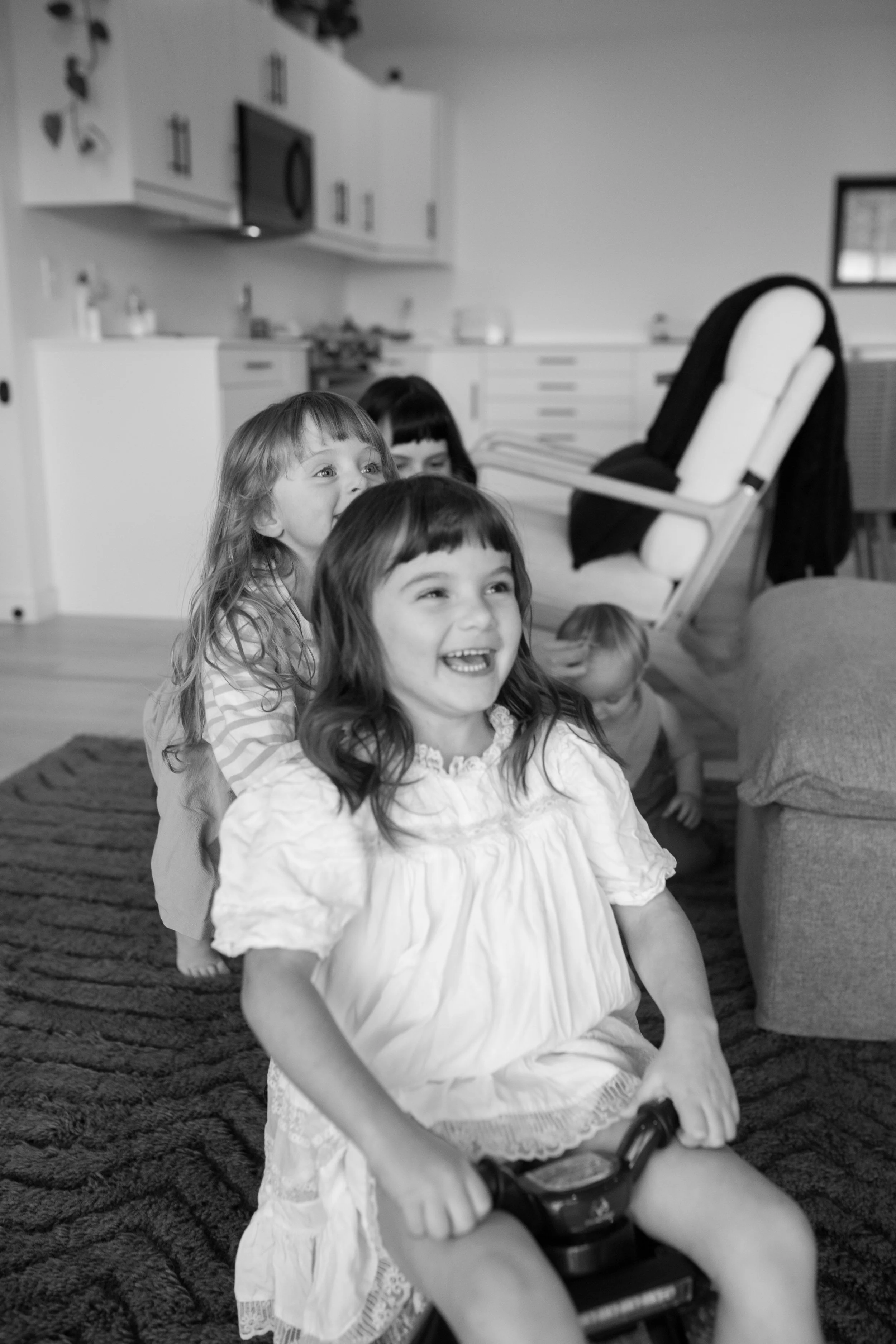 Four young children playing together indoors, with one girl riding a tricycle while the others follow behind, in a living room setting.