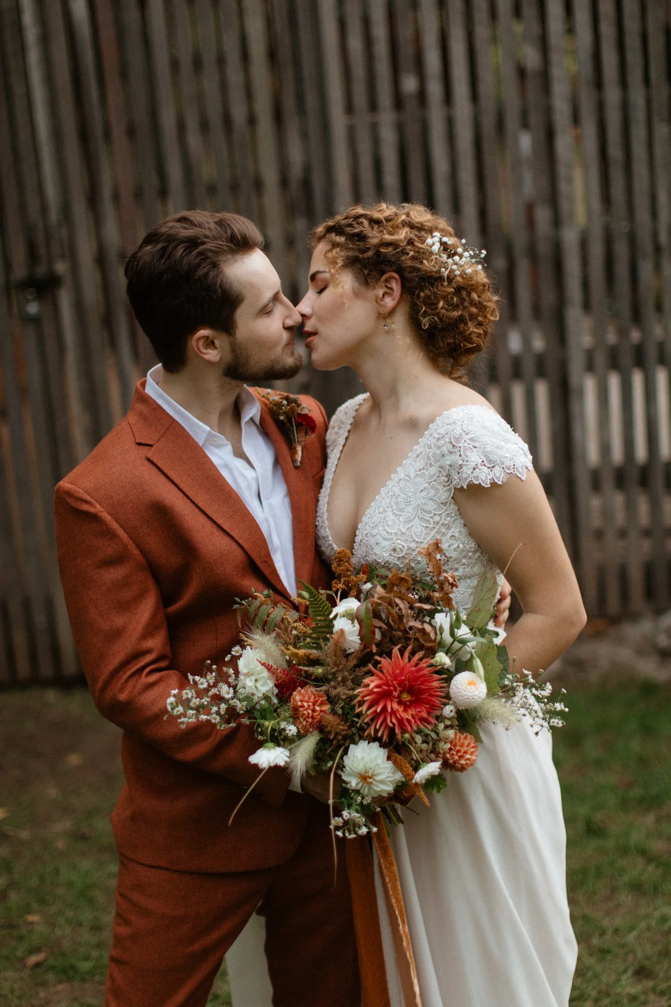 A bride and groom sharing a kiss outdoors, the bride holding a bouquet of flowers and dressed in a lace wedding gown. The groom is wearing a rust-colored suit with a white shirt, with a wooden fence in the background.