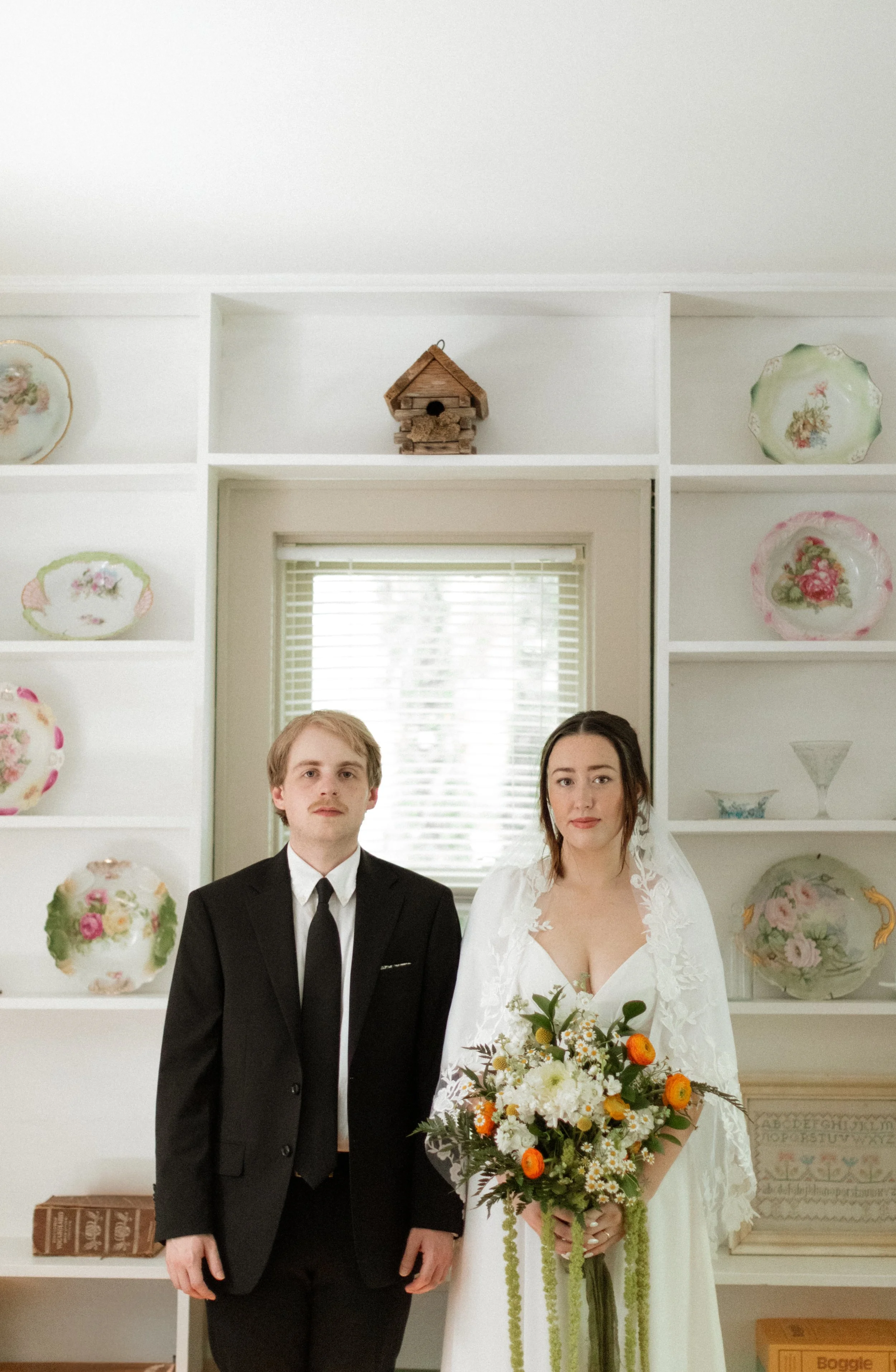 A bride and groom standing in front of a window with white blinds in a white room decorated with floral plates on the shelves. The bride is holding a bouquet and wearing a white dress with lace details, while the groom is dressed in a black suit and 