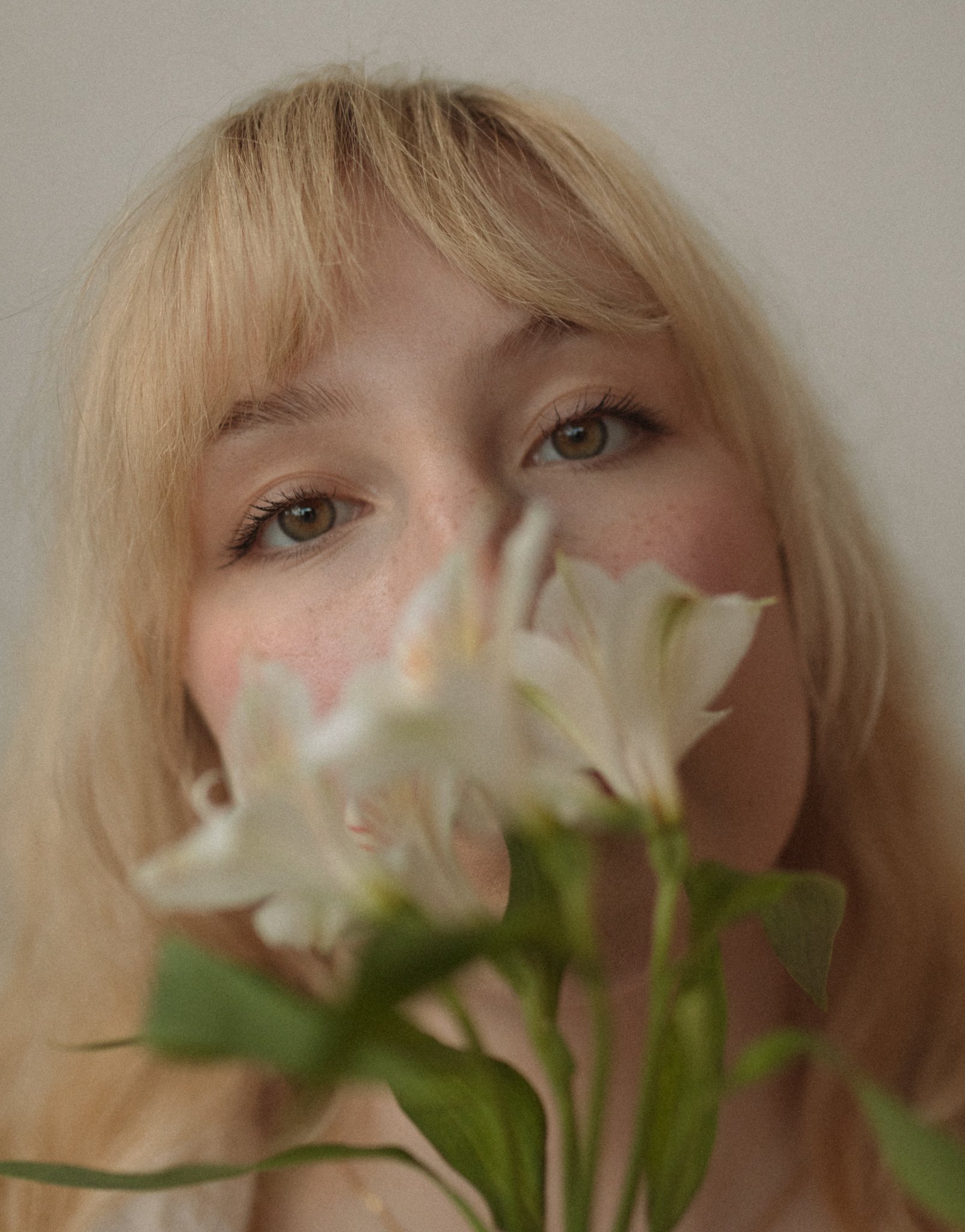 A close-up of a woman with blonde hair and hazel eyes holding white flowers close to her face, partially obscuring her mouth.