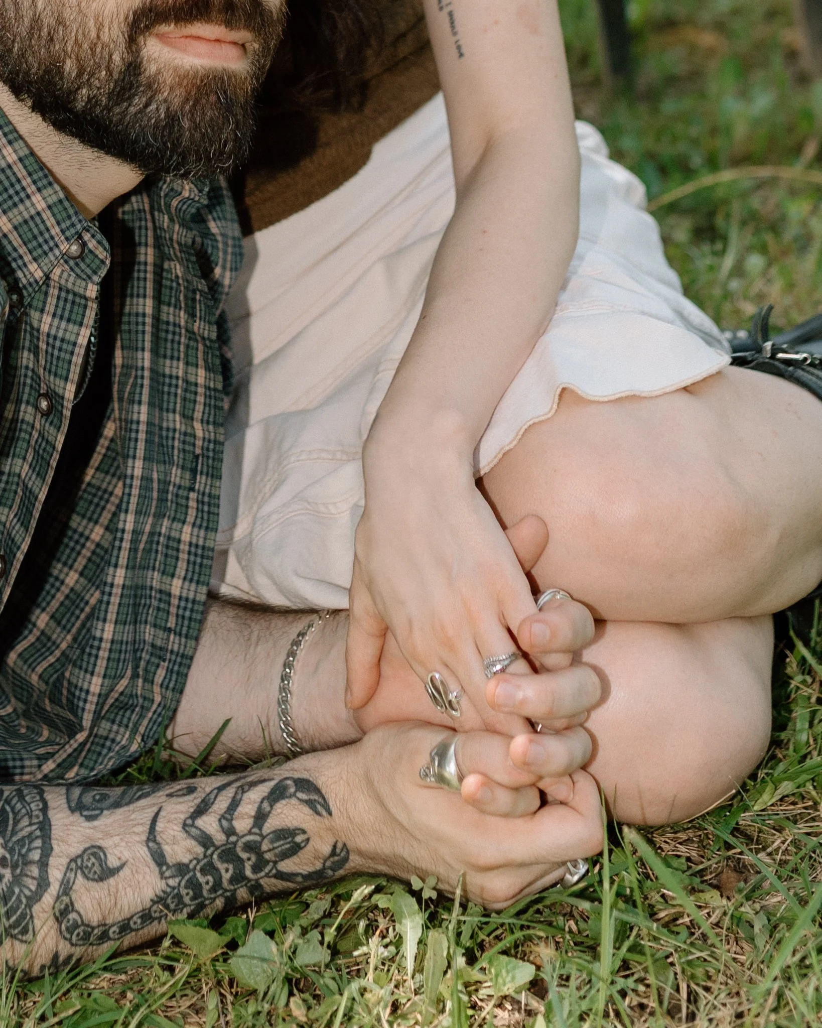 Close-up of two people holding hands while lying on grass, with one person's arm around the other's knee. The person on the left appears to have tattoos on their arm and is wearing rings and a bracelet. The person on the right is wearing a light-colo
