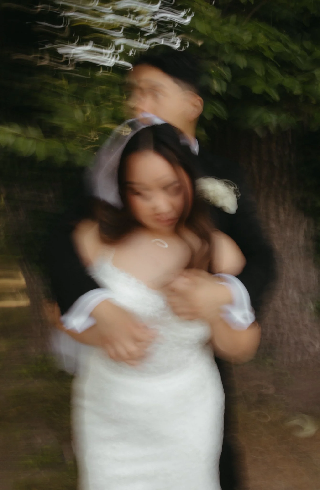 A blurry photo of a couple in formal attire, embracing outdoors with greenery in the background. The woman is in a white dress, and the man is in a dark suit.