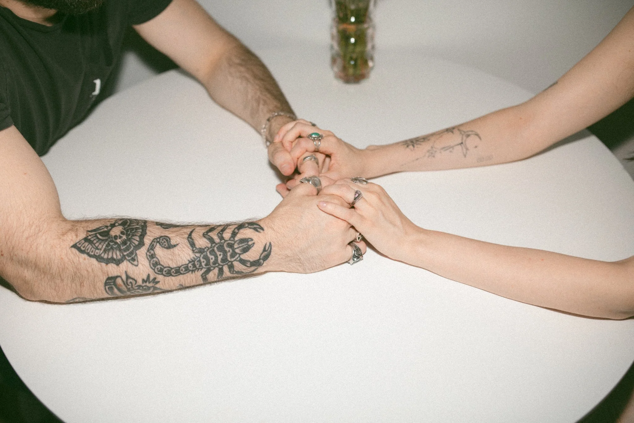Two couples' hands clasped on a white table, with tattoos and rings, and a small vase with flowers in the background.
