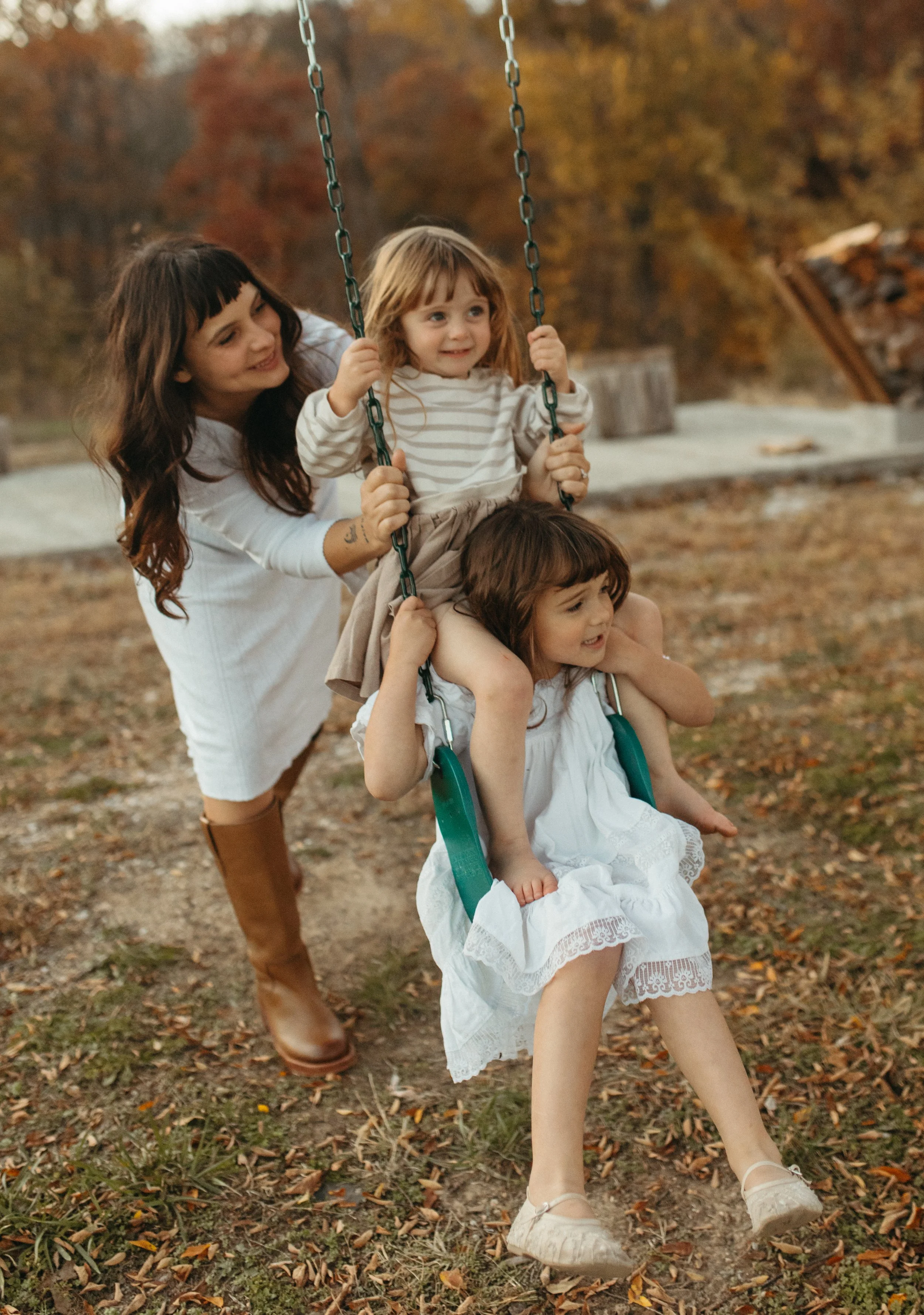 Two young girls sitting on a swing in a park with autumn leaves, accompanied by an older girl pushing the swing.