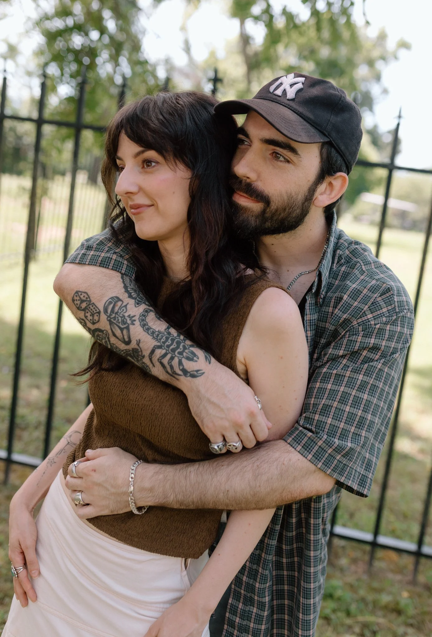 A couple hugging outdoors near a black metal fence, with trees and a house in the background. The woman has dark hair and tattoos on her arm; the man is wearing a checkered shirt and a black baseball cap.