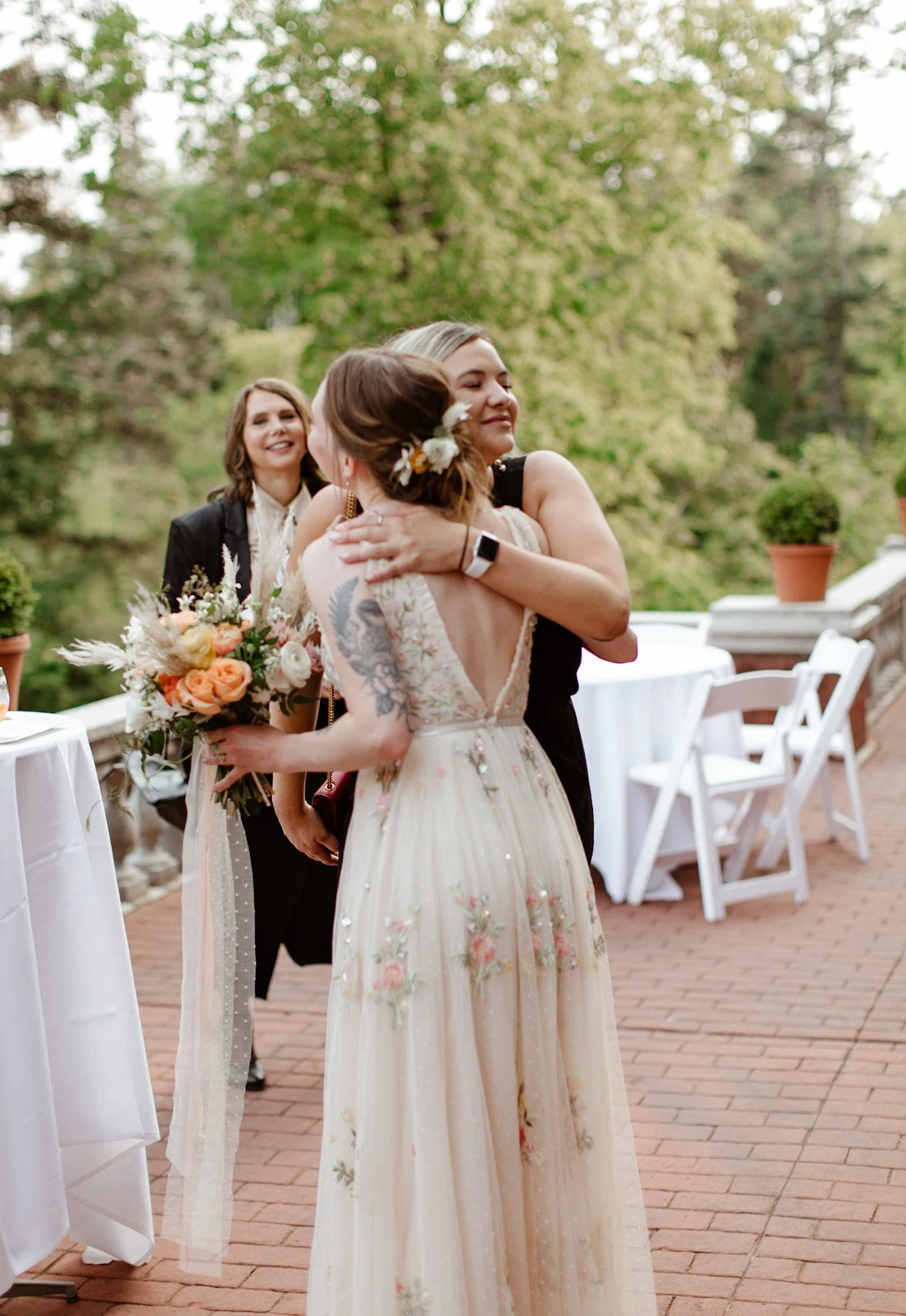 A woman in a black dress hugs another woman in a cream-colored lace dress with floral embroidery, holding a bouquet of flowers, during an outdoor celebration, with another woman smiling in the background.