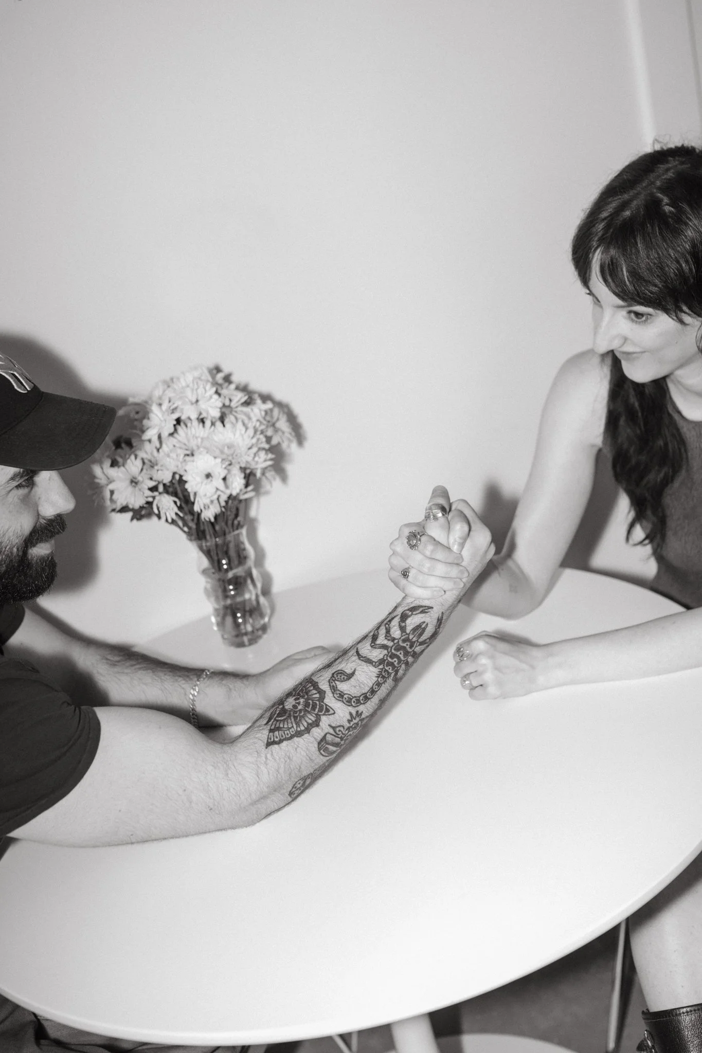 A black and white photo of a man and a woman arm wrestling at a table. The man has a beard and a tattooed arm, wearing a cap, and the woman has long hair and painted nails. A vase of flowers is on the table.