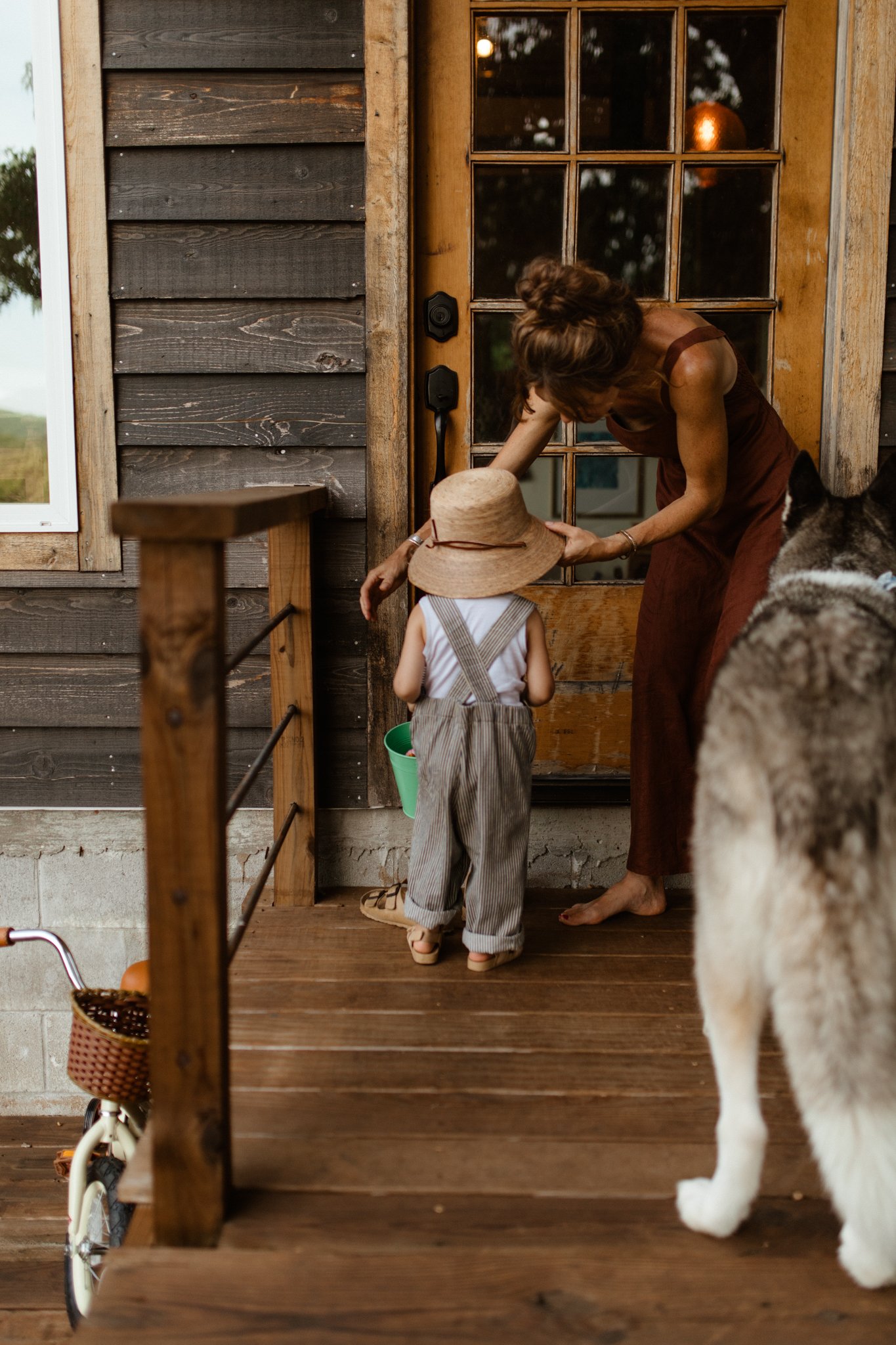 A woman and a young child standing on a porch near a wooden door, with a dog in the foreground. The woman is bending down to talk to the child, who is wearing a large sun hat and striped overalls, holding a small bucket.