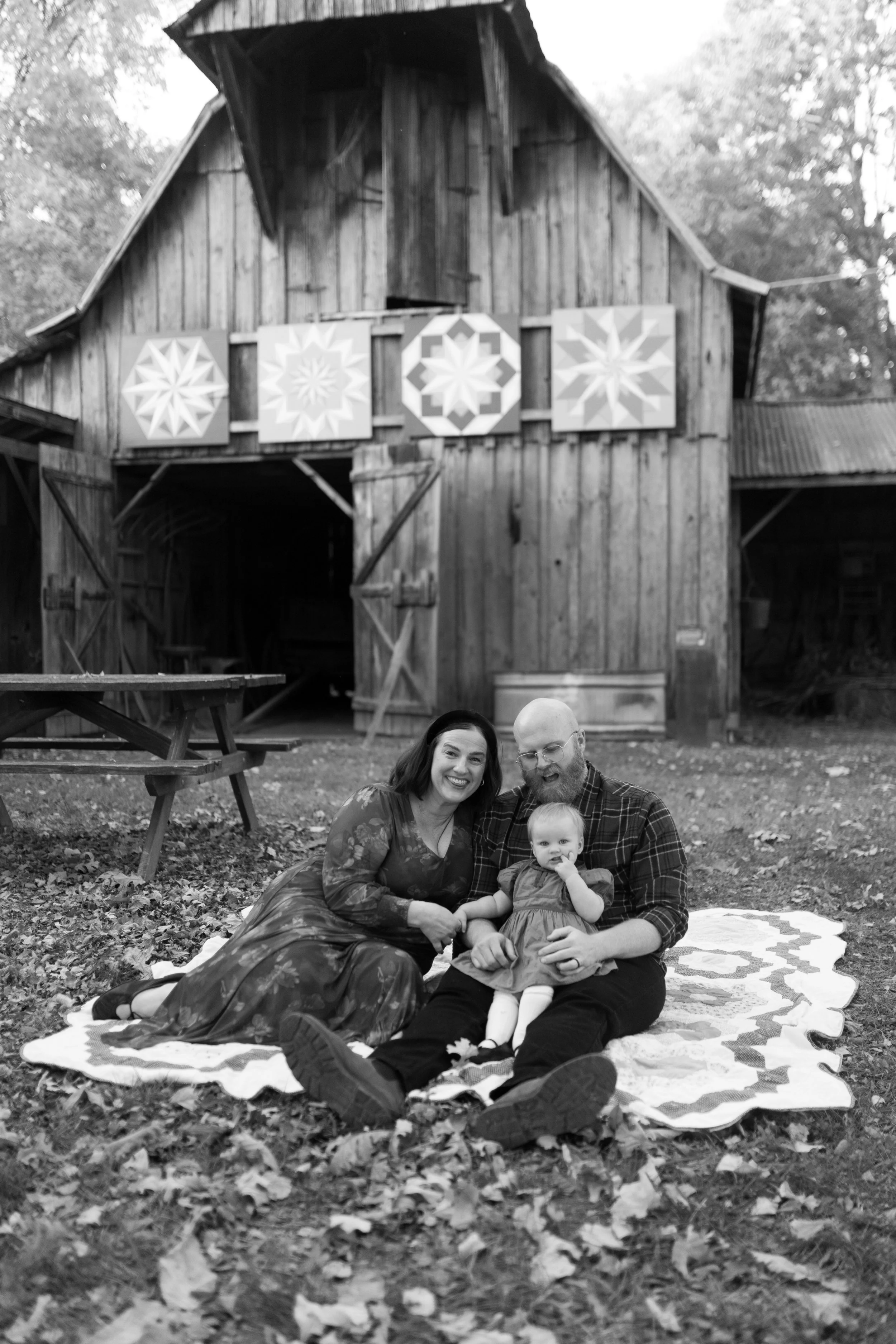 A black and white photo of a family of three sitting on a quilt outdoors, with a wooden barn in the background decorated with quilt patterns, on a fall day with fallen leaves.