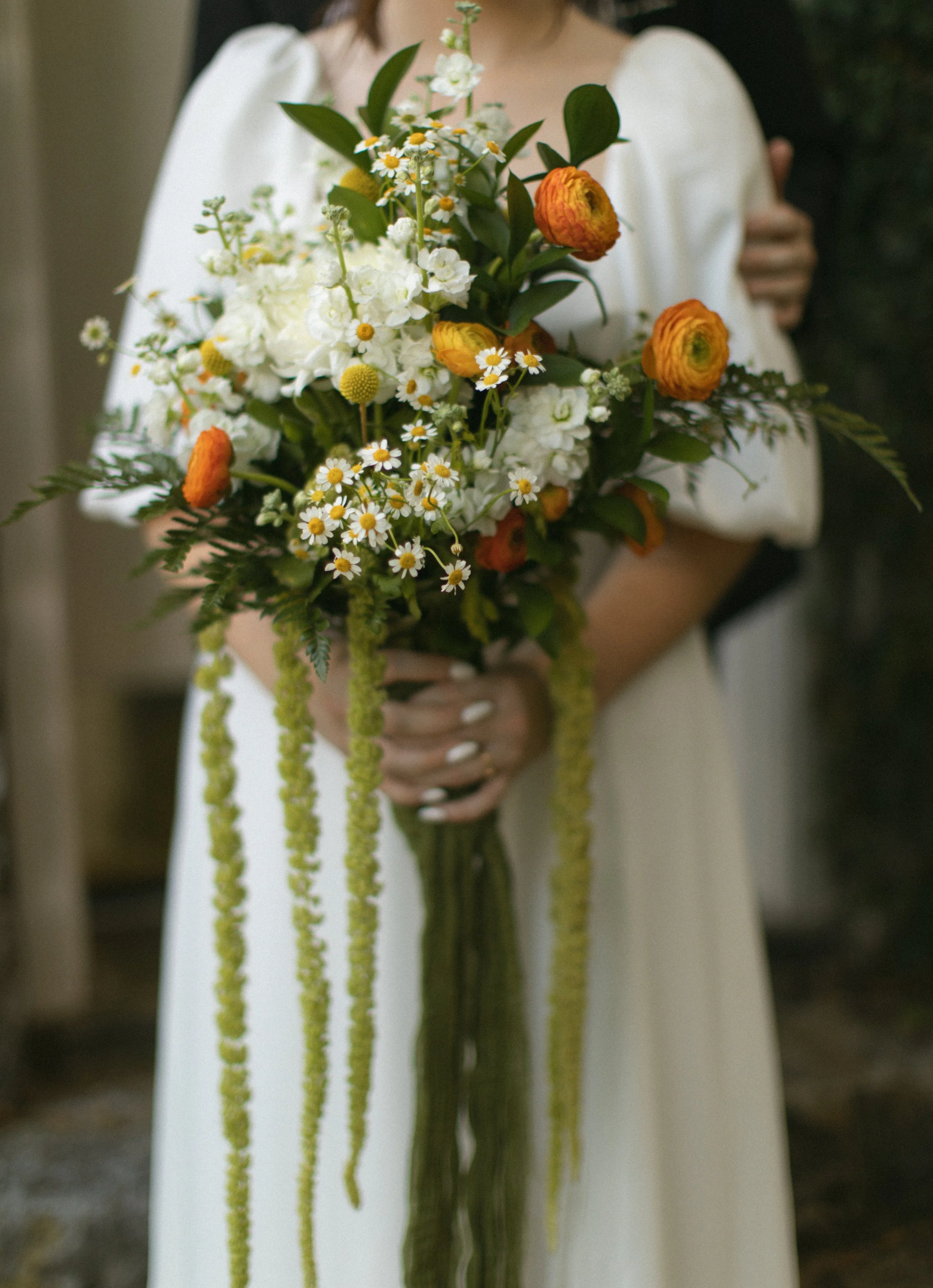 Person in a white dress holding a vibrant bouquet of white, orange, and yellow flowers with greenery.