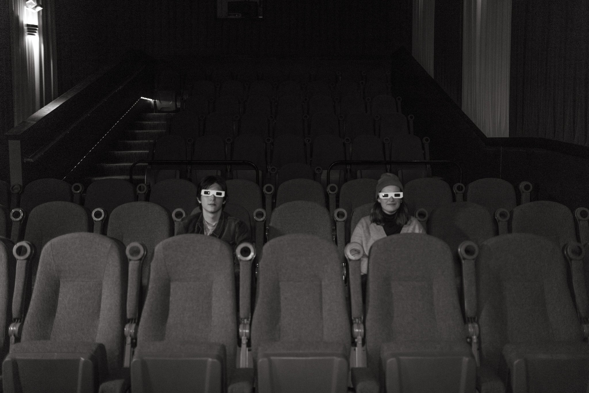 A couple sitting in an empty movie theater wearing 3D glasses, the theater is dark and mostly empty.