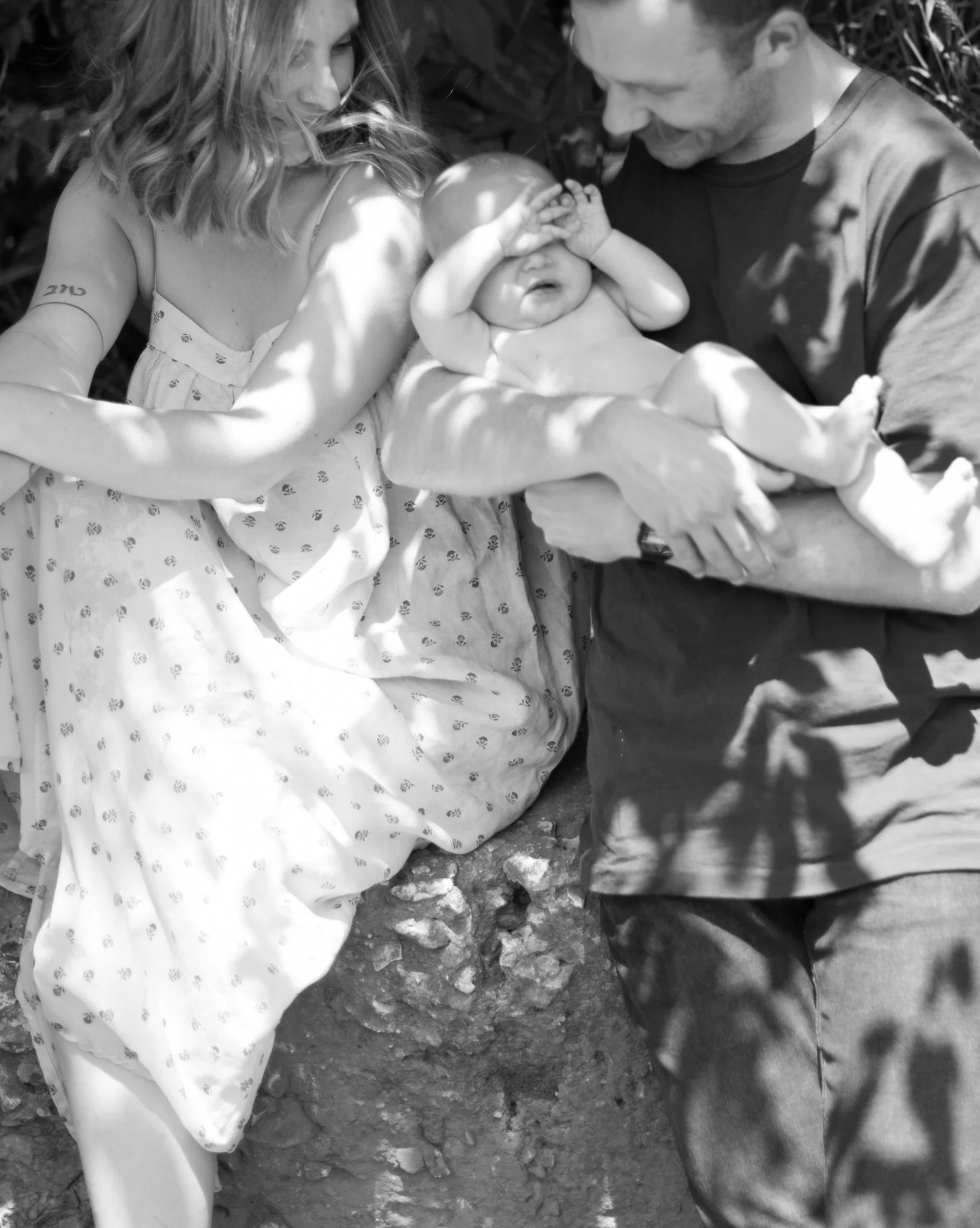A woman and a man holding a baby outdoors, with the woman sitting on a rock and the man standing, both looking at the baby with affection. The woman has shoulder-length wavy hair and a tattoo on her upper arm, and the baby is covering their eyes with