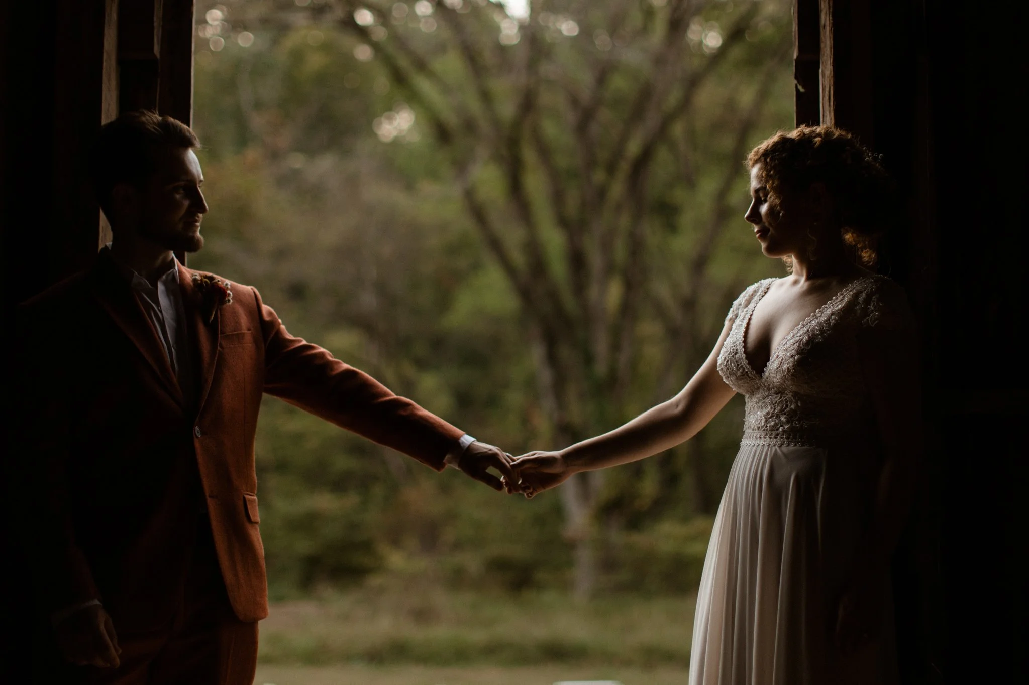 A bride and groom holding hands and facing each other against a natural outdoor background with trees, with the bride in a white wedding dress and the groom in an orange suit.